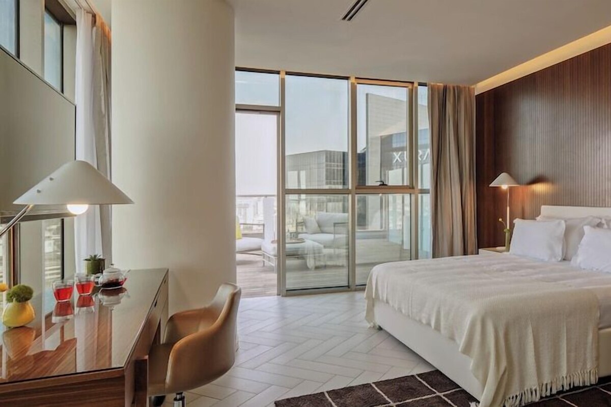 A hotel room with a large bed, a desk with a lamp, a chair, and a view of the outside through a glass door.