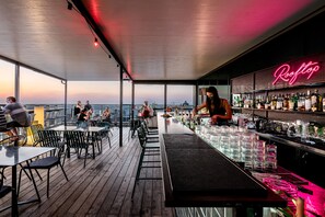 A rooftop bar with a neon sign, wooden deck, and tables and chairs for guests. - Prima Millennium (Ra'ananna)