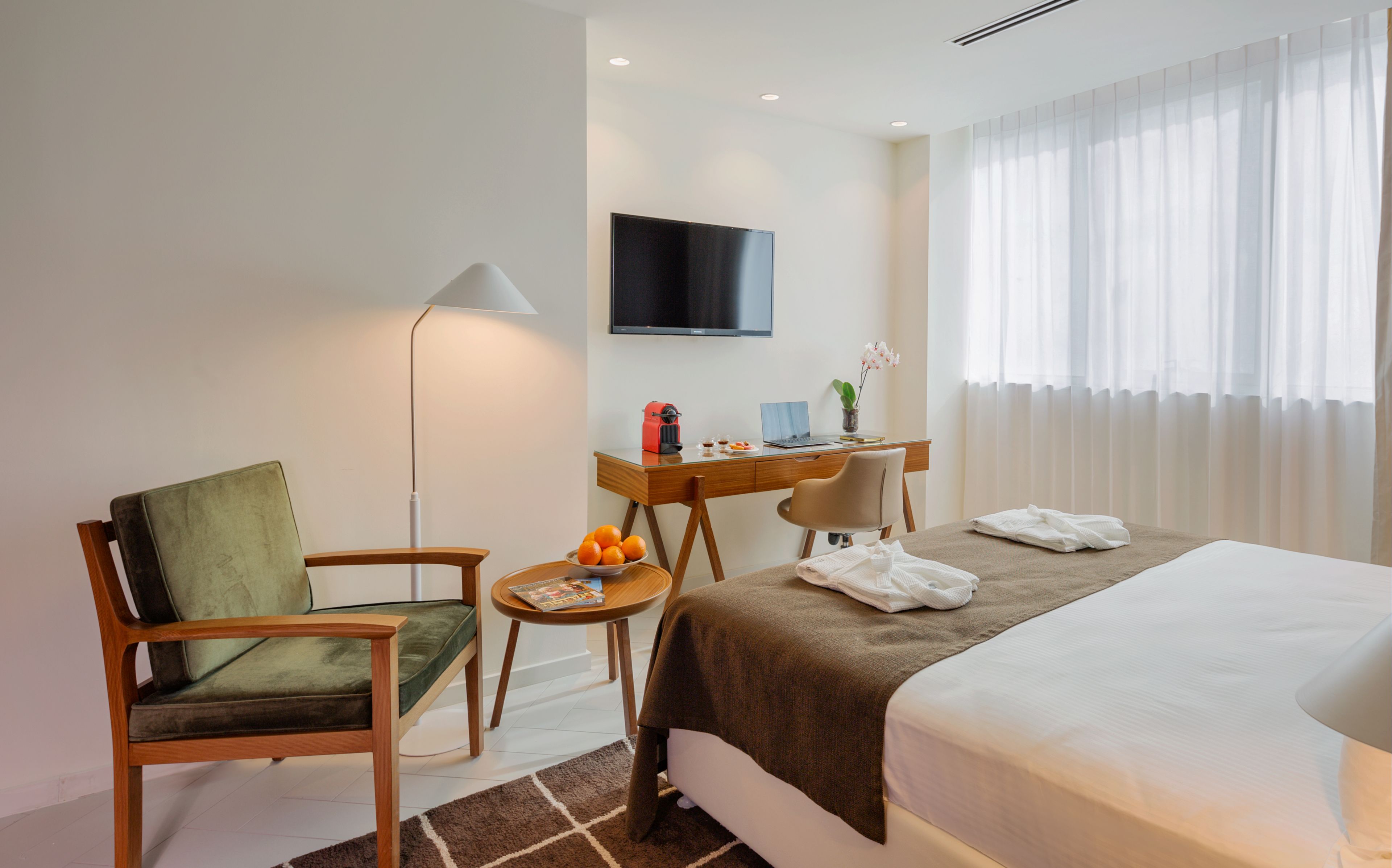 Quarto de hotel com uma cama, uma escrivaninha com um laptop, uma cadeira, um abajur e uma TV na parede.