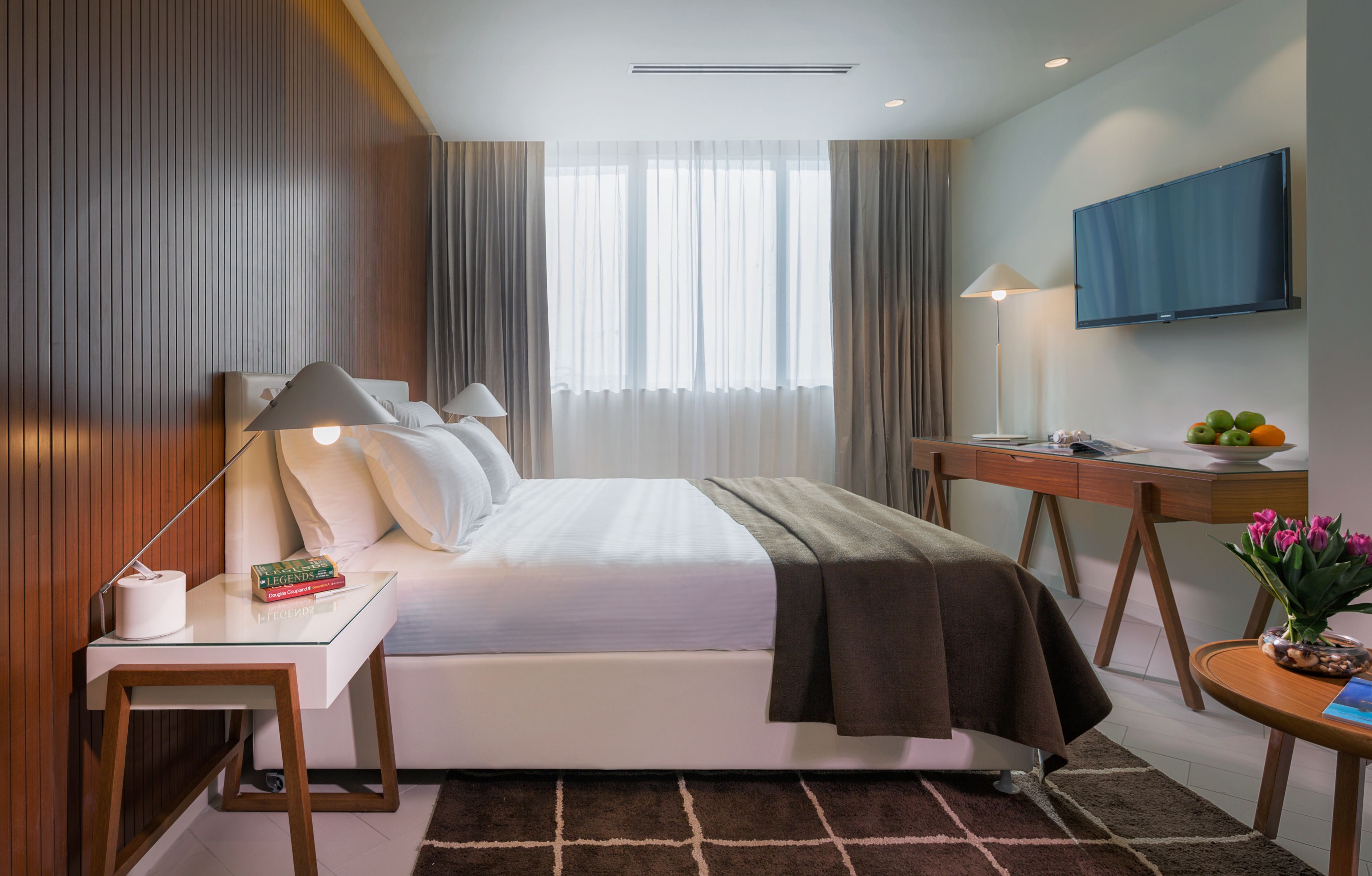 A modern hotel room with a neatly made bed, a desk with a lamp, a flat-screen TV, and a small table with fruit and flowers.