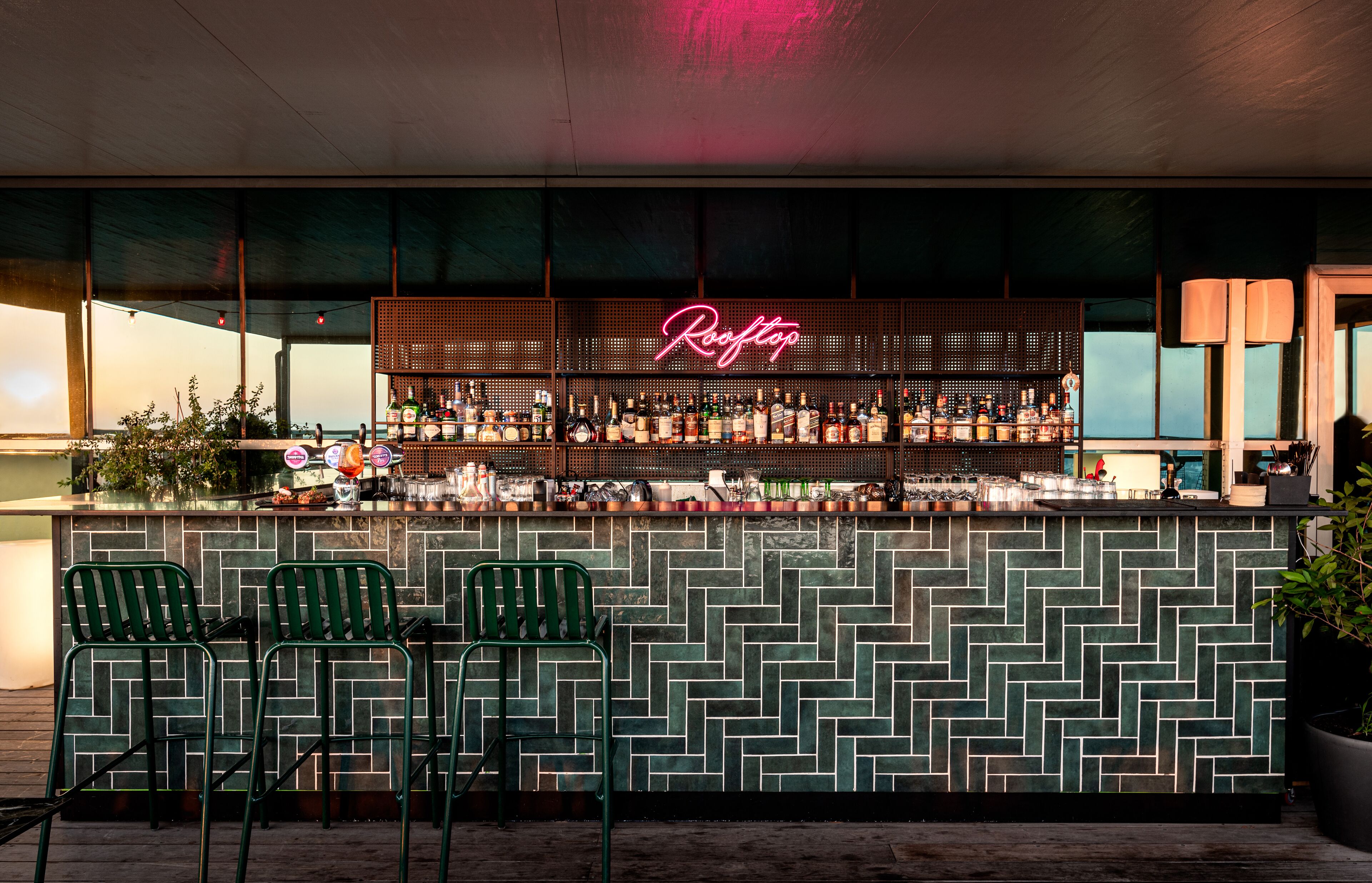 Zona de bar con un letrero de neón, sillas verdes y una pared de azulejos.