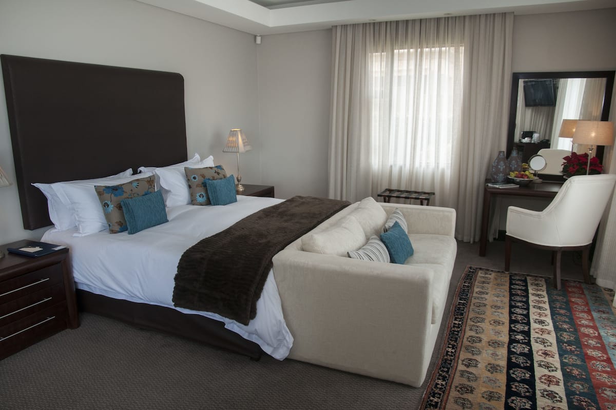 A hotel room with a large bed, a sofa, a chair, a desk with a lamp, and a mirror.