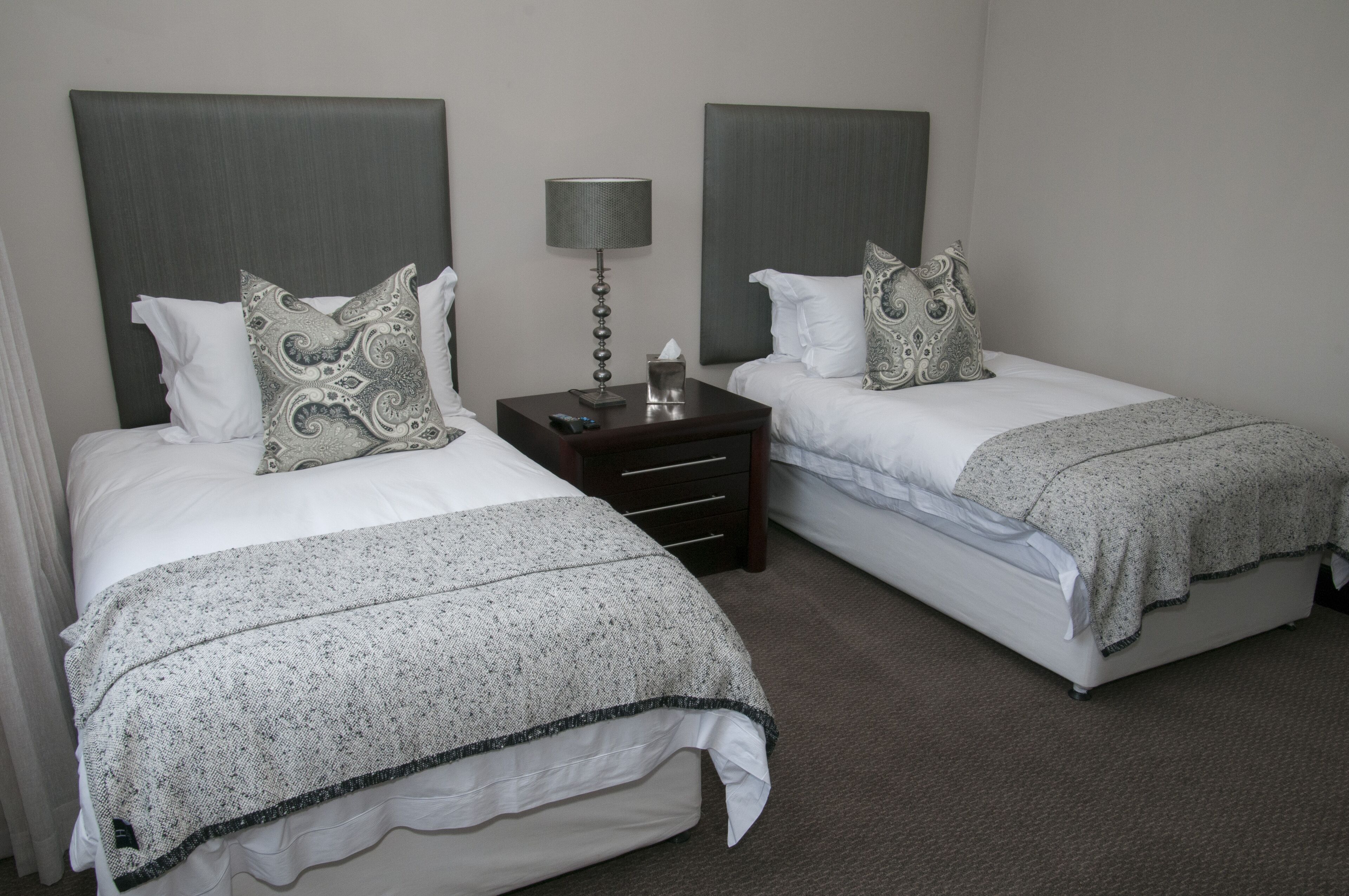 Une chambre d’hôtel avec deux lits simples, chacun doté d’une tête de lit grise, de draps blancs et d’oreillers décoratifs. On y trouve une table de chevet avec une lampe et une boîte de mouchoirs.