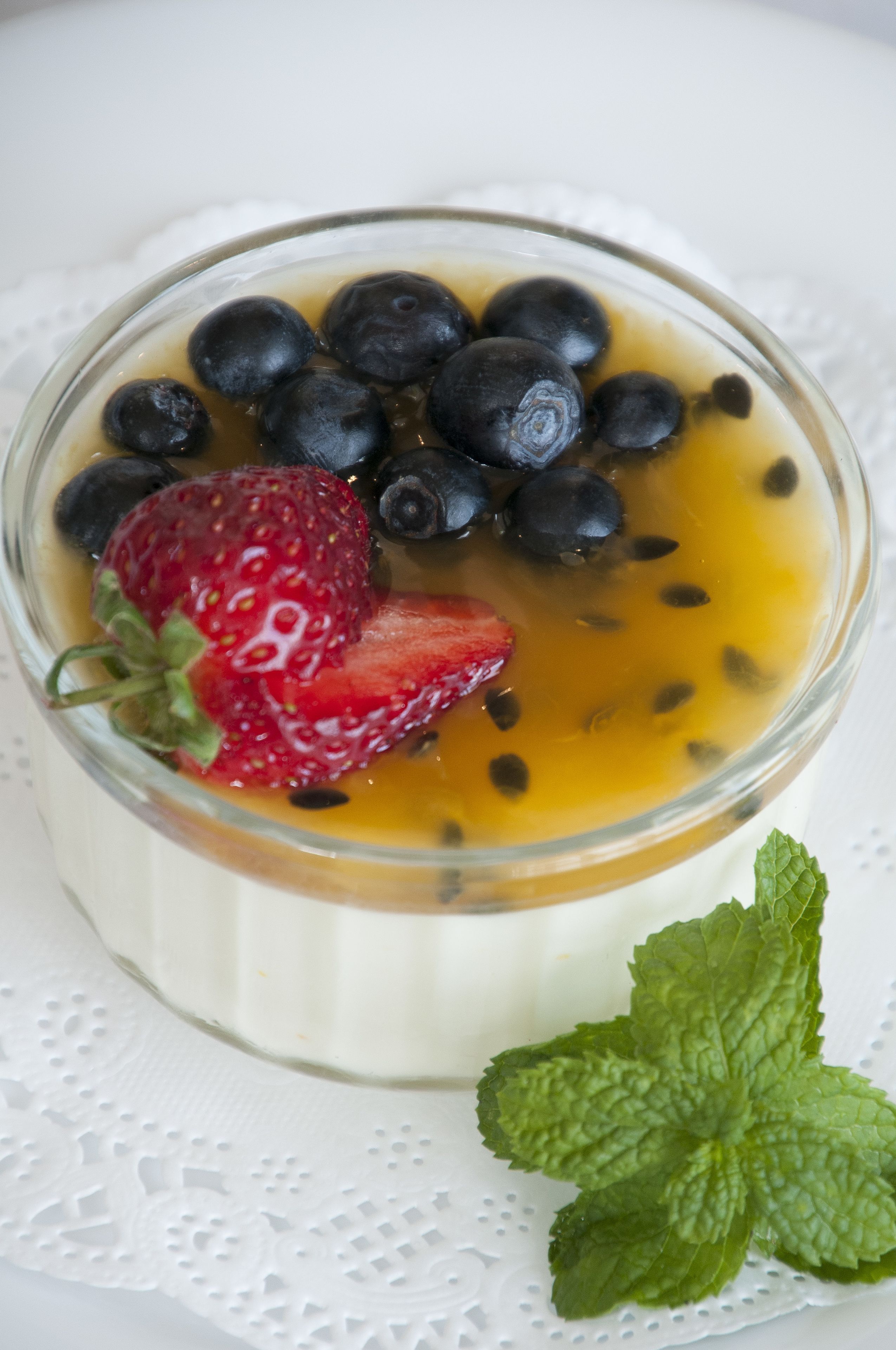 Postre en un cuenco de vidrio transparente con fresas y arándanos por encima, sobre un encaje blanco.