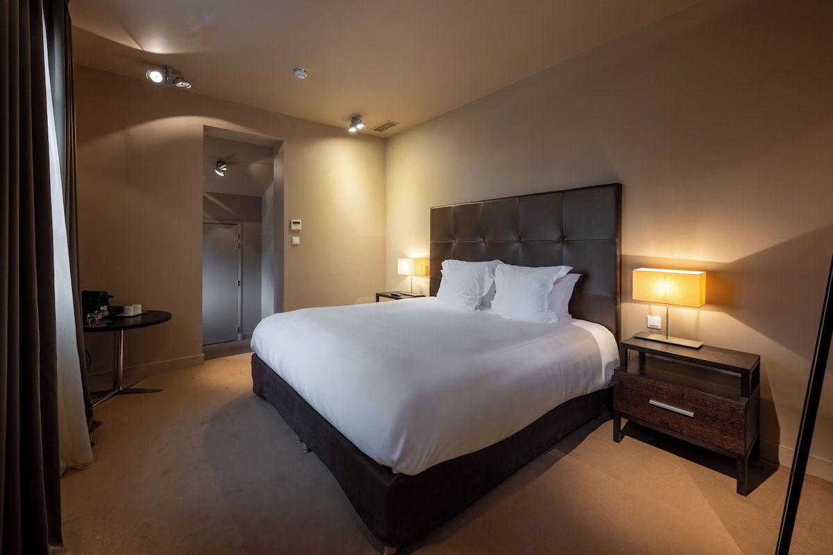 Ein Hotelzimmer mit einem großen Bett, Nachttischlampen, einem kleinen Tisch und einer Tür zu einem anderen Raum.