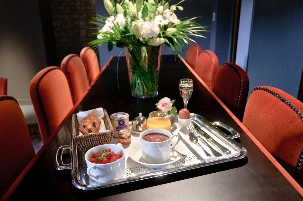 A breakfast tray with croissants, a bowl of strawberries, a cup of coffee, a glass of orange juice, a chocolate dessert, and a bottle of milk on a table with a vase of flowers and a bottle of champagne.