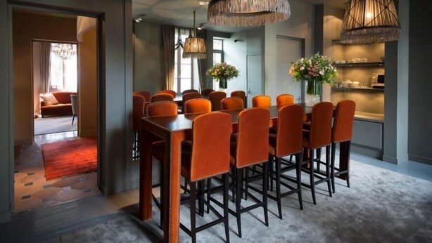 A dining area with a long table and orange chairs, two hanging lamps, and a view into another room with a red carpet. - Hotel Ligaro - Jarnac (Jarnac)