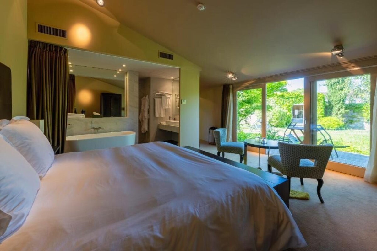Ein Hotelzimmer mit einem großen Bett, einer Sitzecke mit Tisch und Stühlen, einem Badezimmer mit Spiegel und Blick auf Grünflächen.