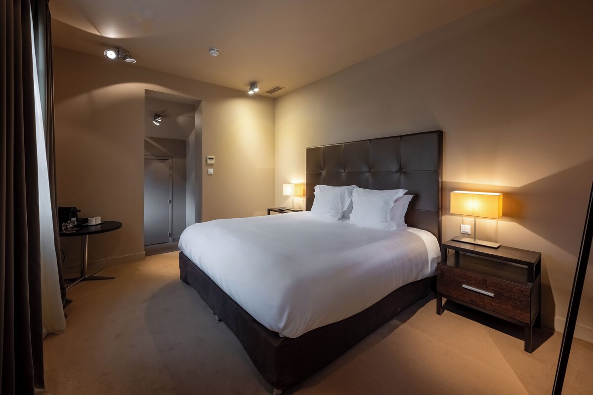 A hotel room with a large bed, bedside lamps, a small table, and a door leading to another room.