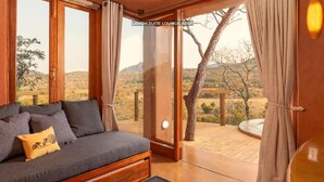A room with a sofa, curtains, and a view of a desert landscape. - Leshiba Wilderness (Louis Trichardt)