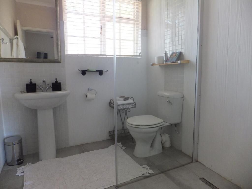 A small, clean bathroom with a white sink, toilet, and a mirror above the sink. There is a window with blinds, a small shelf with toiletries, and a trash can.