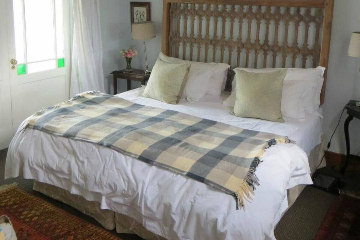 A bedroom with a wooden headboard, a bed with white bedding and a plaid blanket, a nightstand with a lamp, and a window with curtains.
