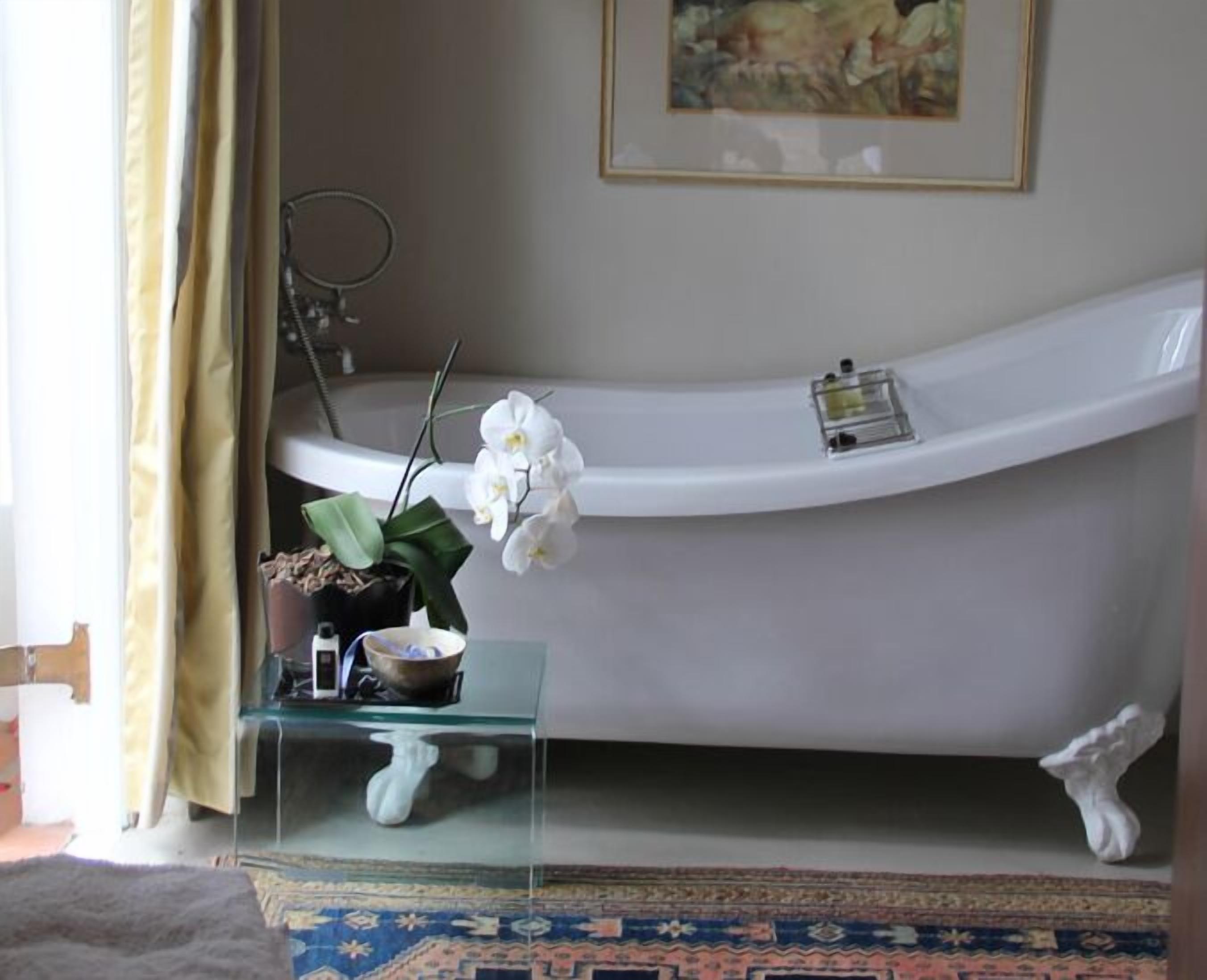 Une salle de bain avec une baignoire, une étagère en verre où sont rangés les articles de toilette, une orchidée en pot et un tableau encadré au mur.