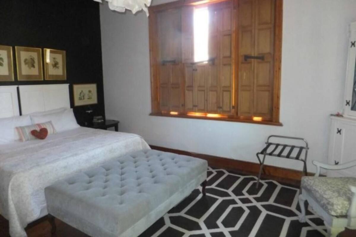 A bedroom with a bed, a bench, a window with wooden shutters, and a patterned rug.