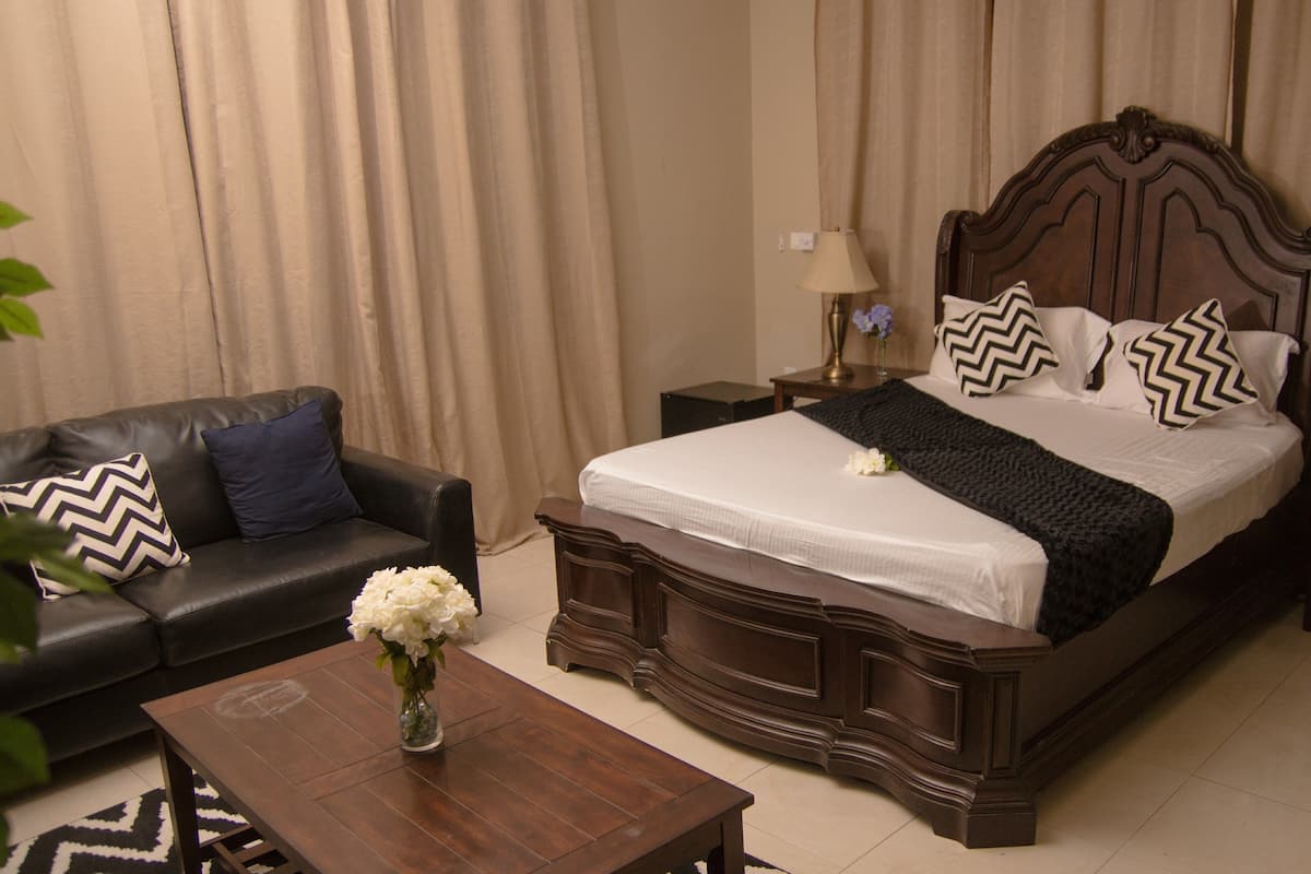 Habitación de hotel con una cama grande, cabecera de madera oscura, sofá de cuero negro, mesita de noche con lámpara y un jarrón con flores blancas sobre una mesa de centro de madera.