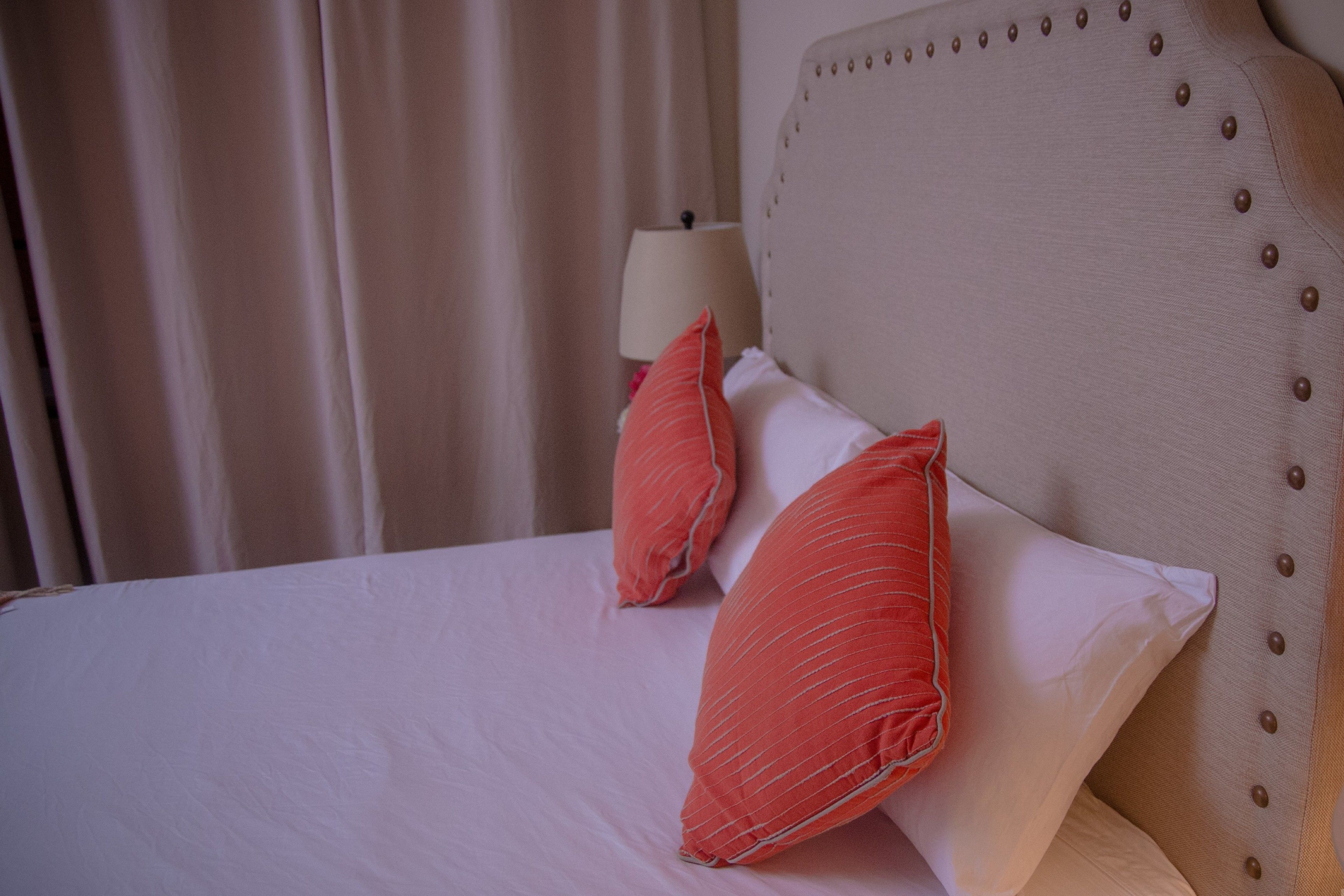 A neatly made bed with white and orange pillows, a beige upholstered headboard with buttons, and a bedside lamp.