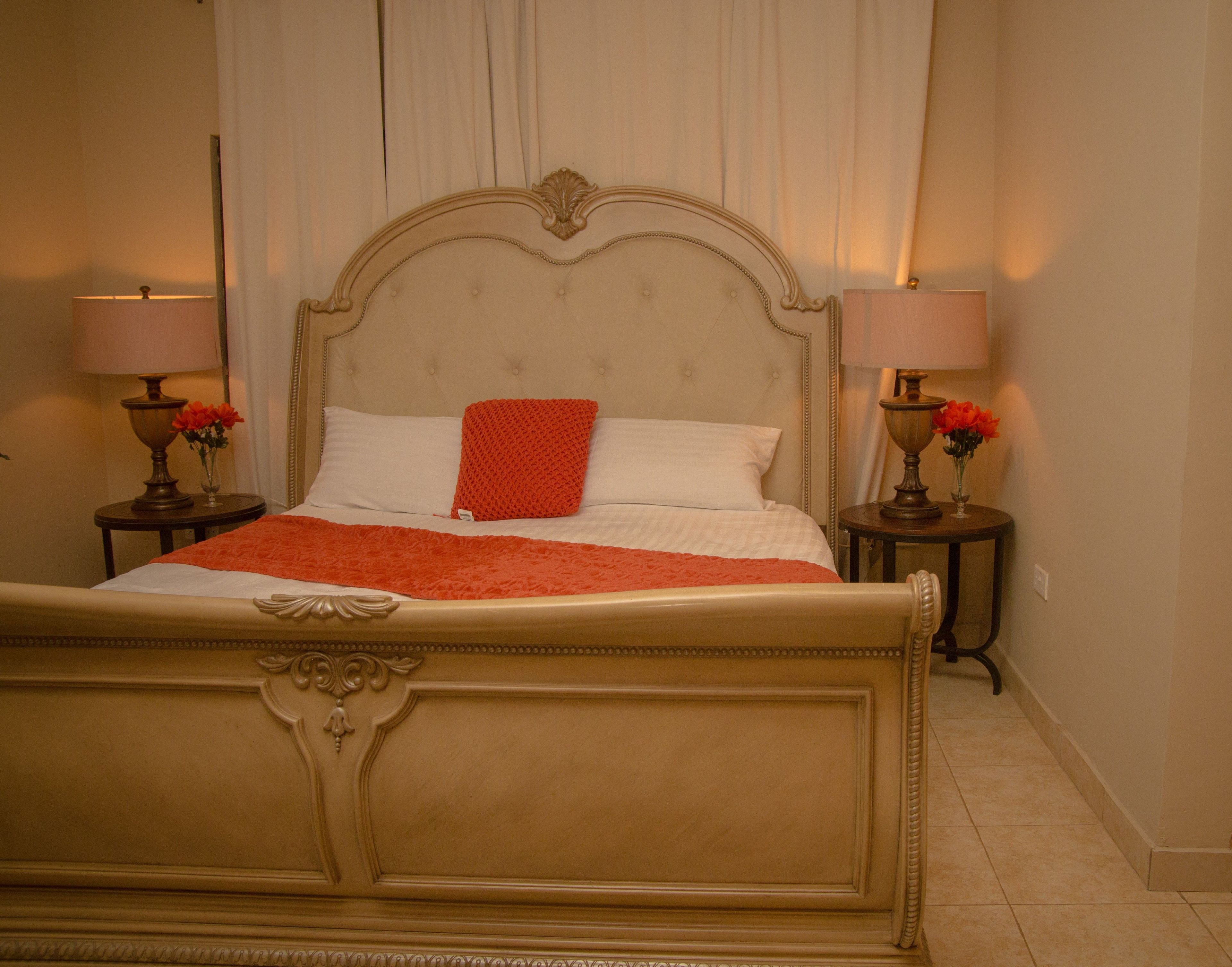 Un lit avec un oreiller rouge, deux tables de chevet avec des lampes et des fleurs, et un rideau en arrière-plan.