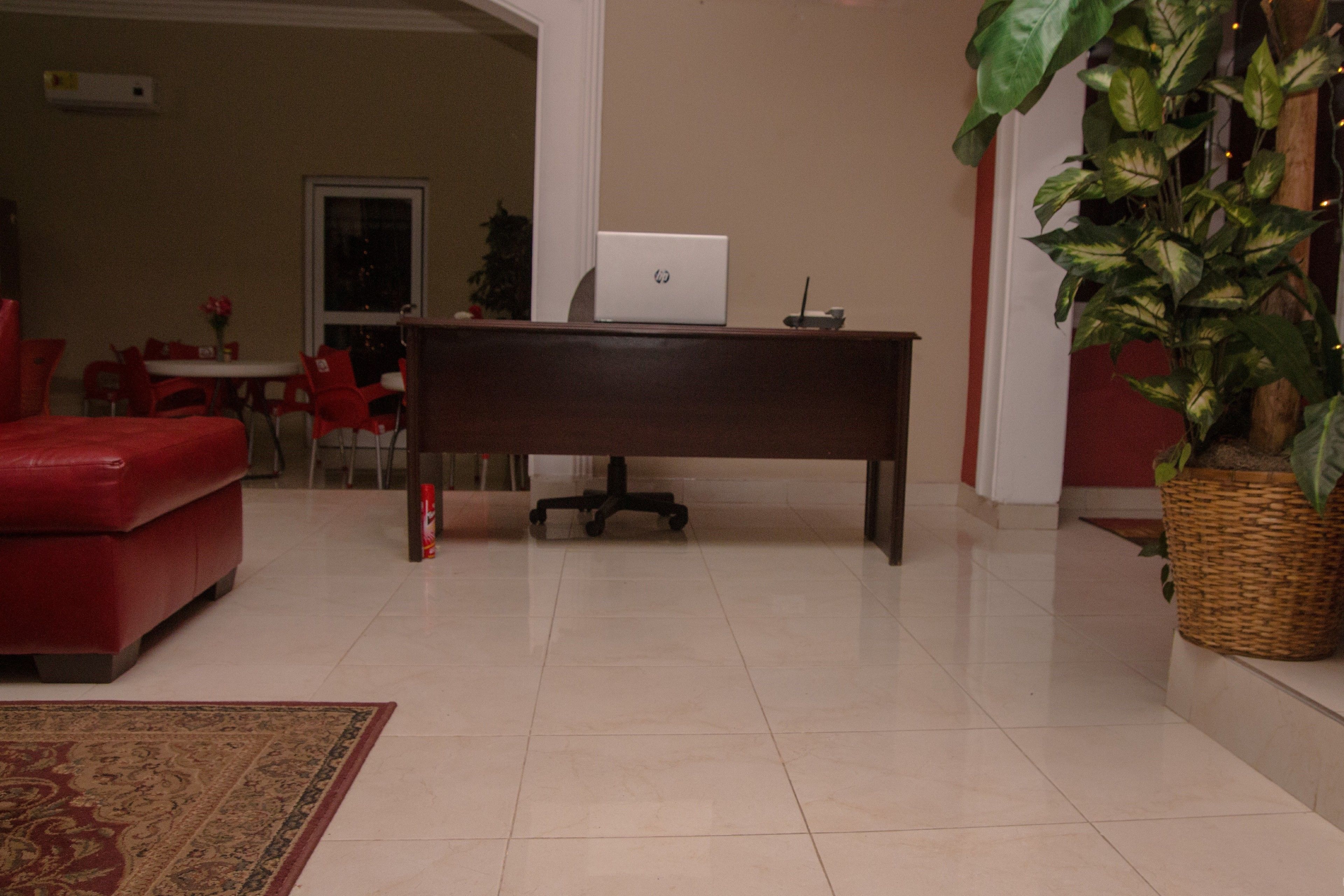 A living room with a red sofa, a wooden desk with a laptop, a red chair, a dining table with chairs, and a potted plant.