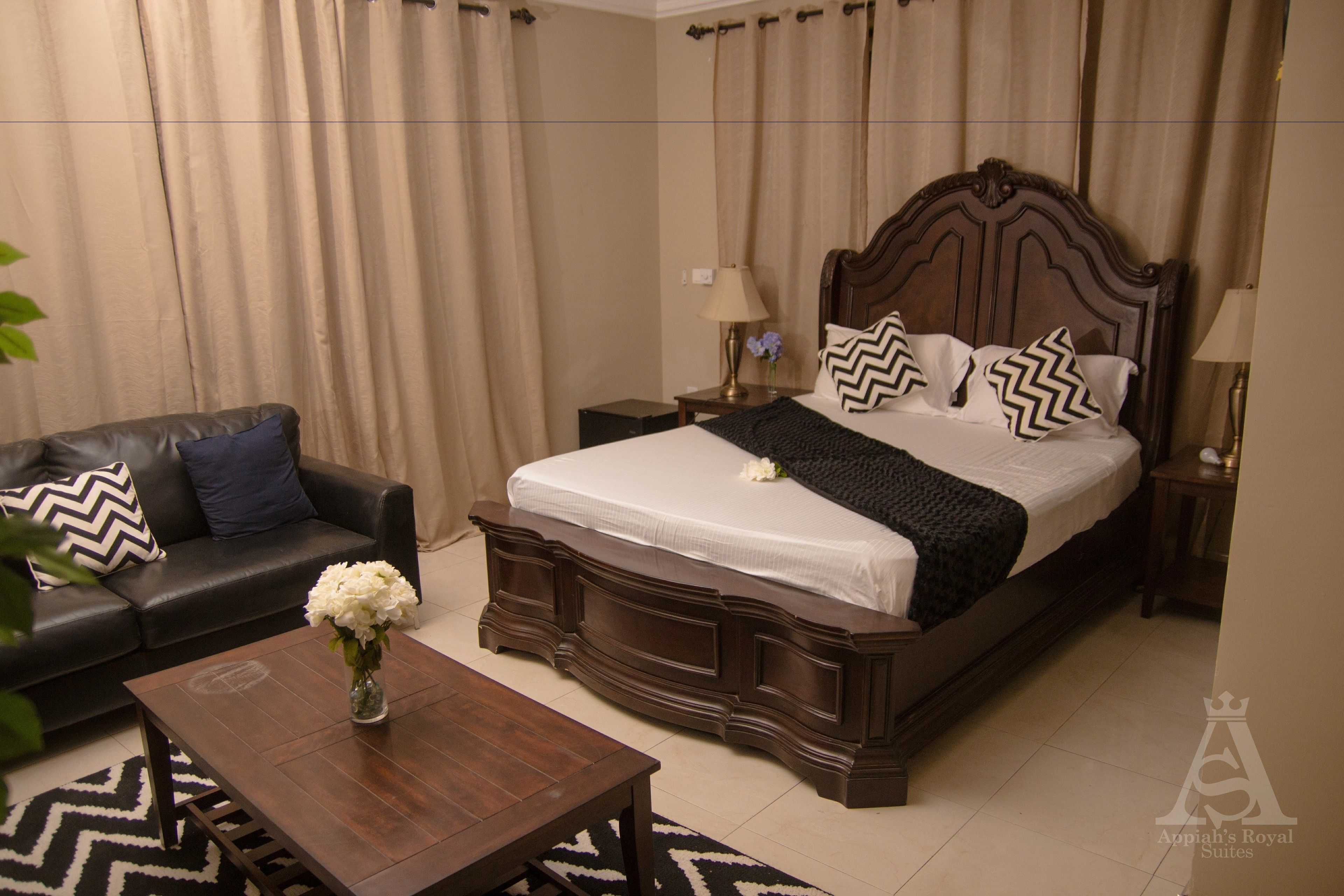A hotel room with a large bed, a dark wooden headboard, a black leather sofa, a wooden coffee table, and a vase with white flowers.