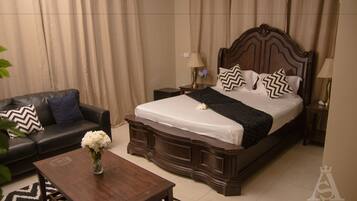 A hotel room with a large bed, a dark wooden headboard, a black leather sofa, a wooden coffee table, and a vase with white flowers.