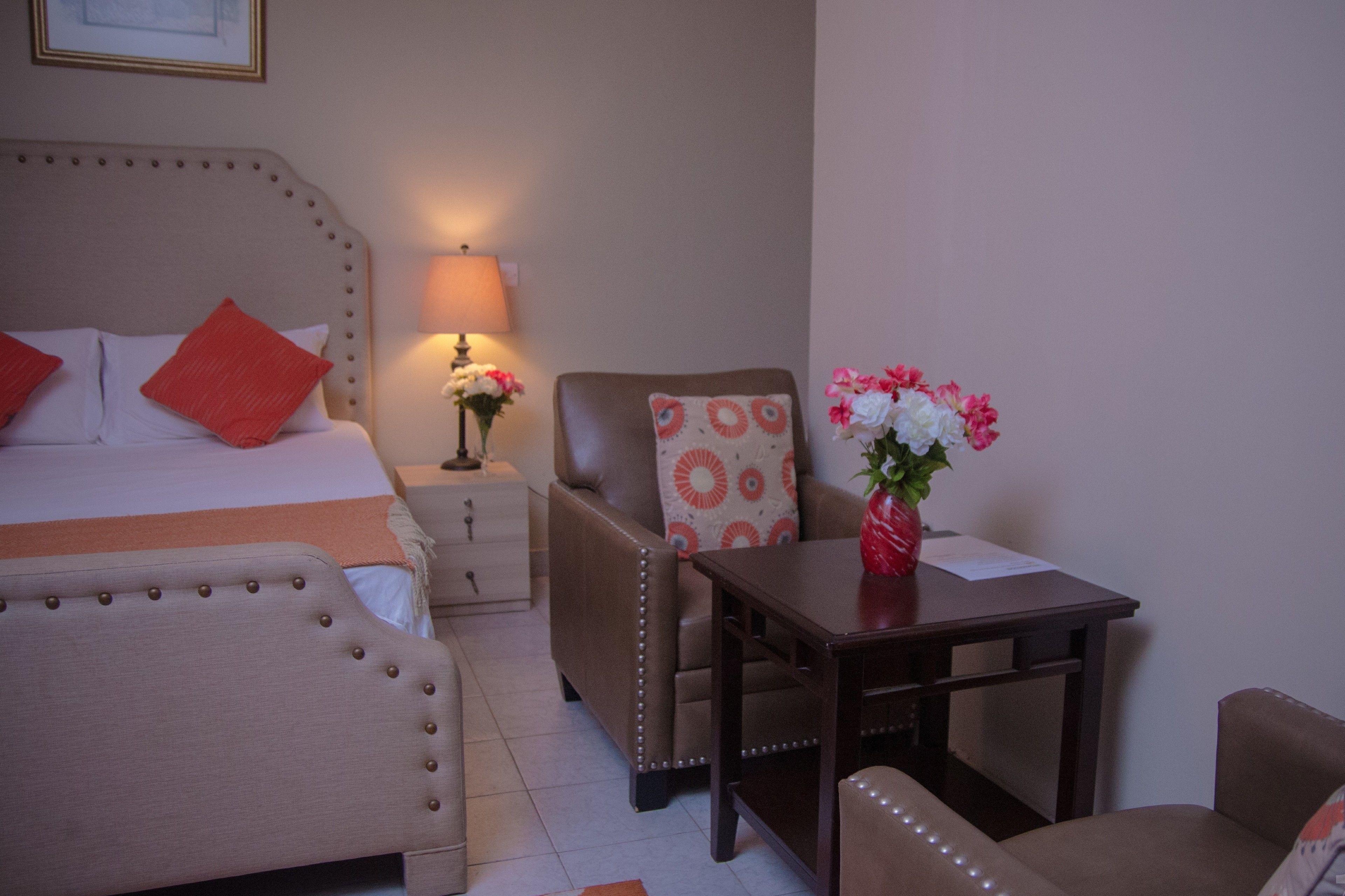Habitación de hotel con cama, dos sillas, una mesita, lámpara y un florero con flores.