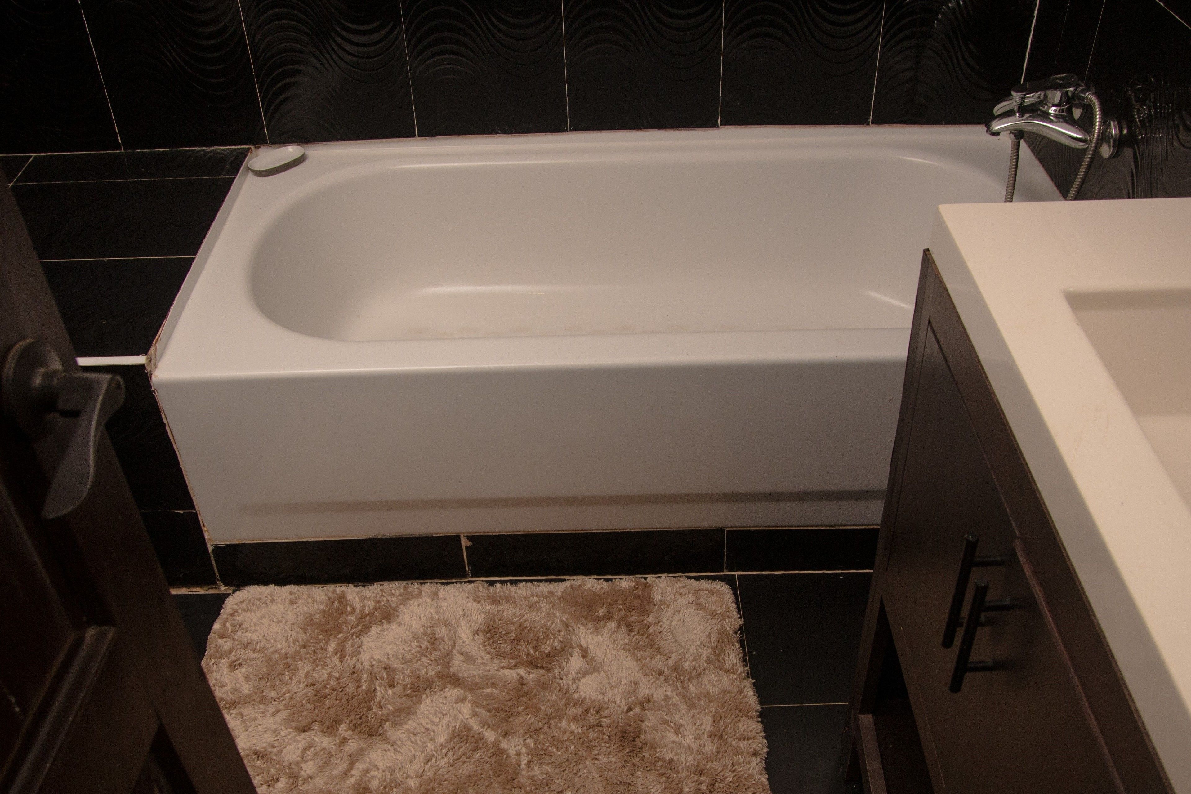 A bathroom with a white bathtub, dark tiled walls, and a fluffy beige bath mat.