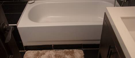 Un baño con una bañera blanca, paredes cubiertas de baldosas oscuras y una alfombra de baño color beige y mullida.