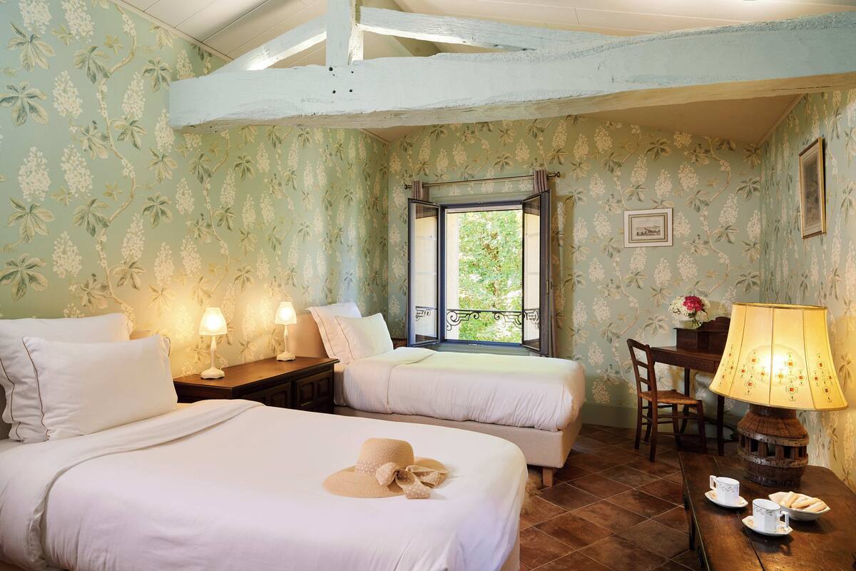 A room with two beds, a table with a lamp, and a window with a view of greenery.