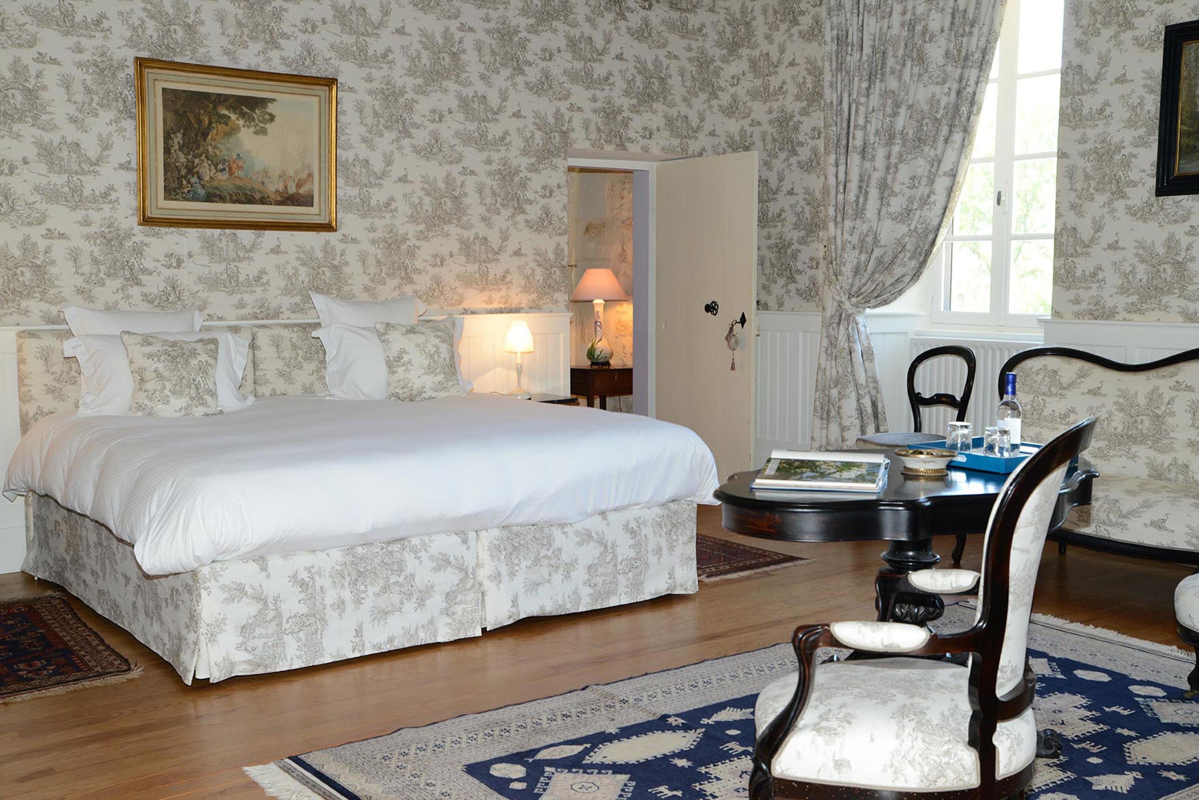 Un dormitorio con una cama grande, una silla, una mesa con libros y una ventana con cortinas.