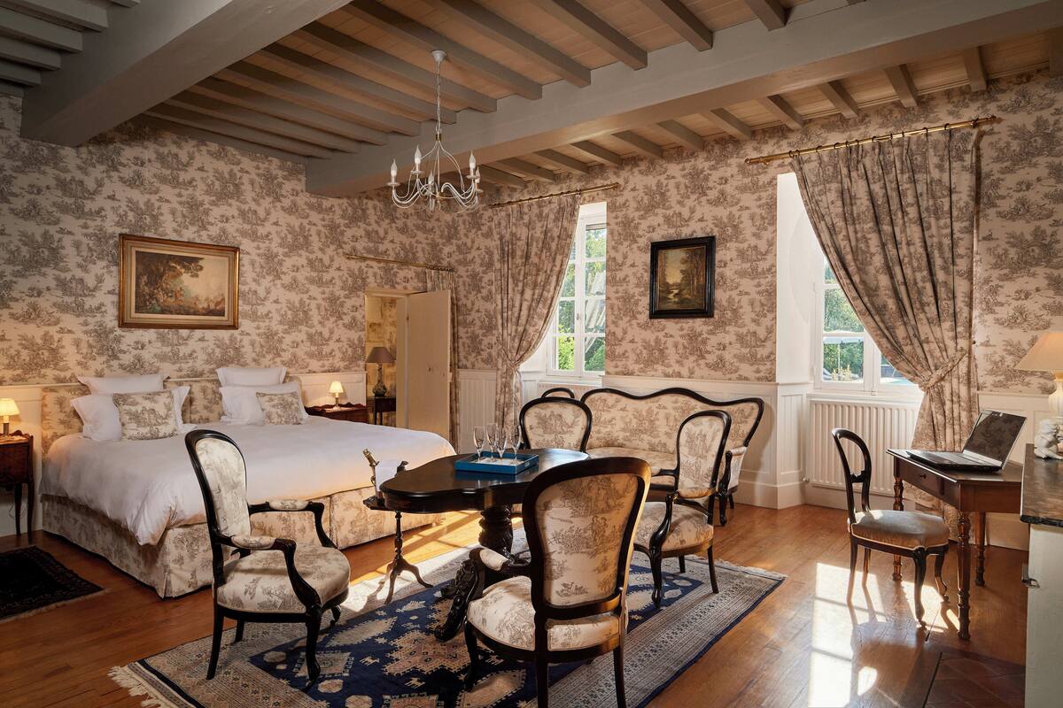 A bedroom with a large bed, a dining table with chairs, a laptop on a desk, and a chandelier.