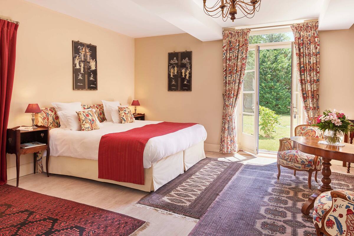 A bedroom with a large bed, a nightstand, a chair, a table with a vase, and a view of the garden through the window.