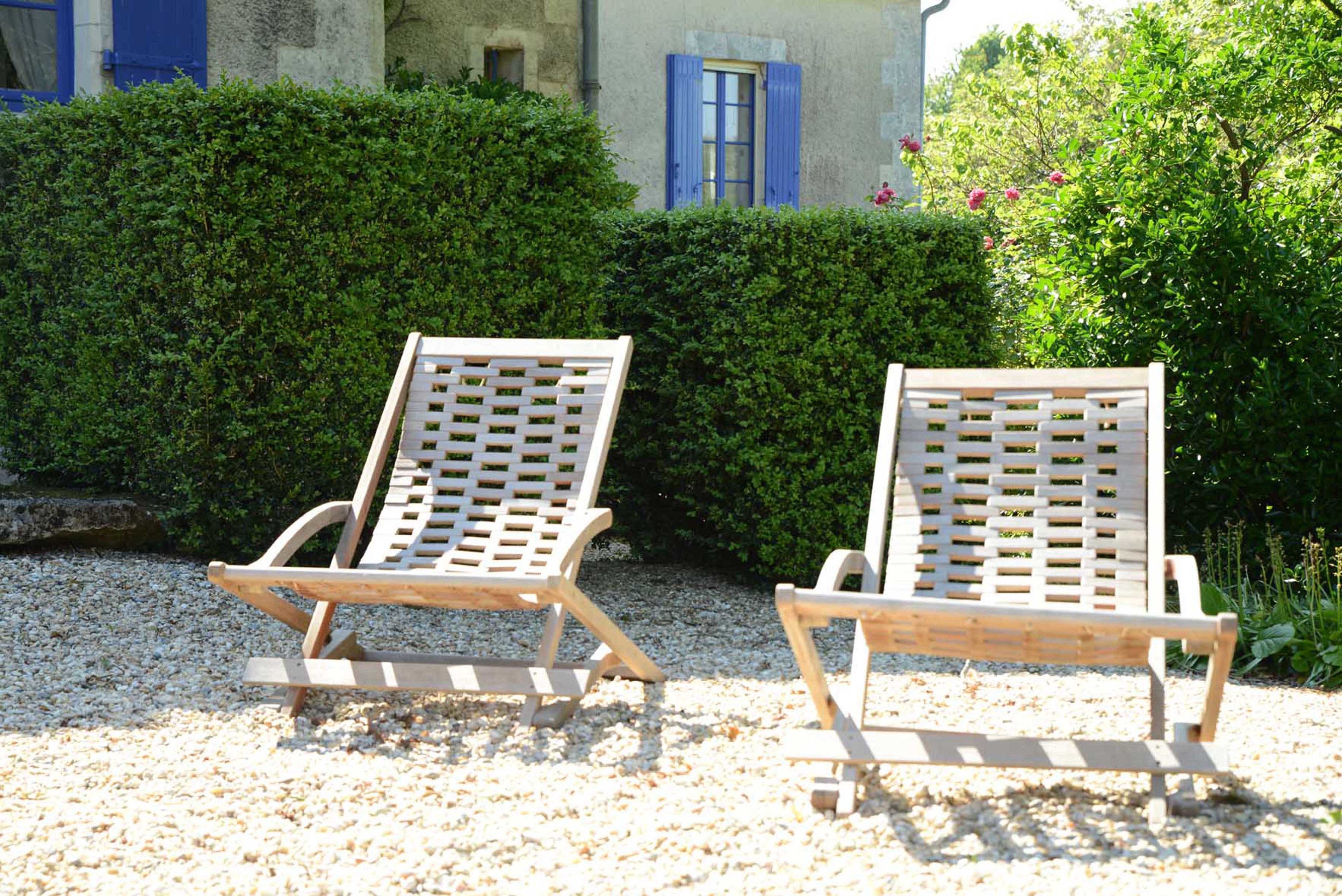 Two wooden lounge chairs with a lattice backrest design, positioned on a gravel surface with a hedge and a building with blue shutters in the background.