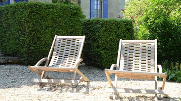 Two wooden lounge chairs with a lattice backrest design, positioned on a gravel surface with a hedge and a building with blue shutters in the background.