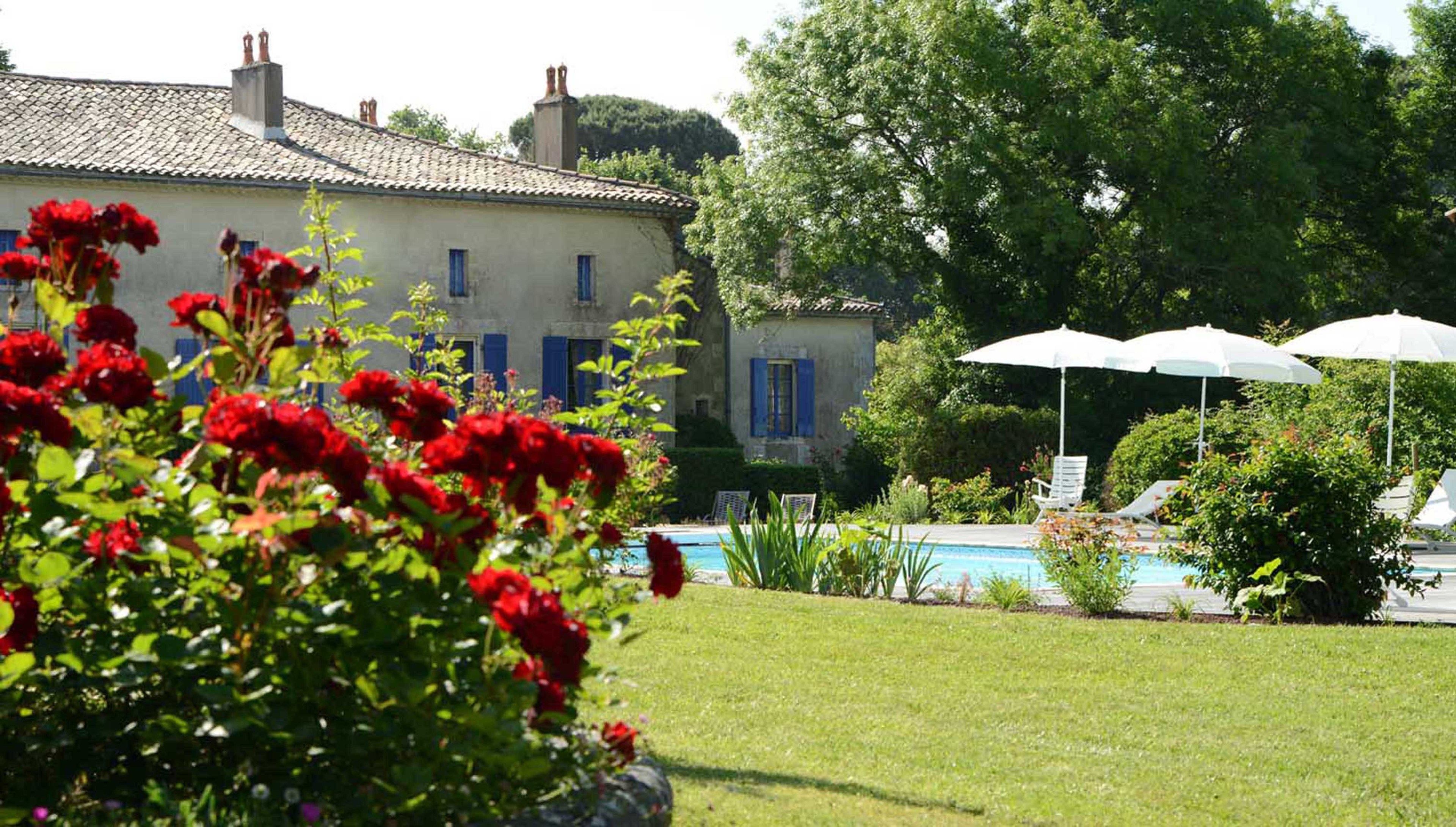 A pool area with lounge chairs and umbrellas, surrounded by lush greenery and a building with blue shutters.