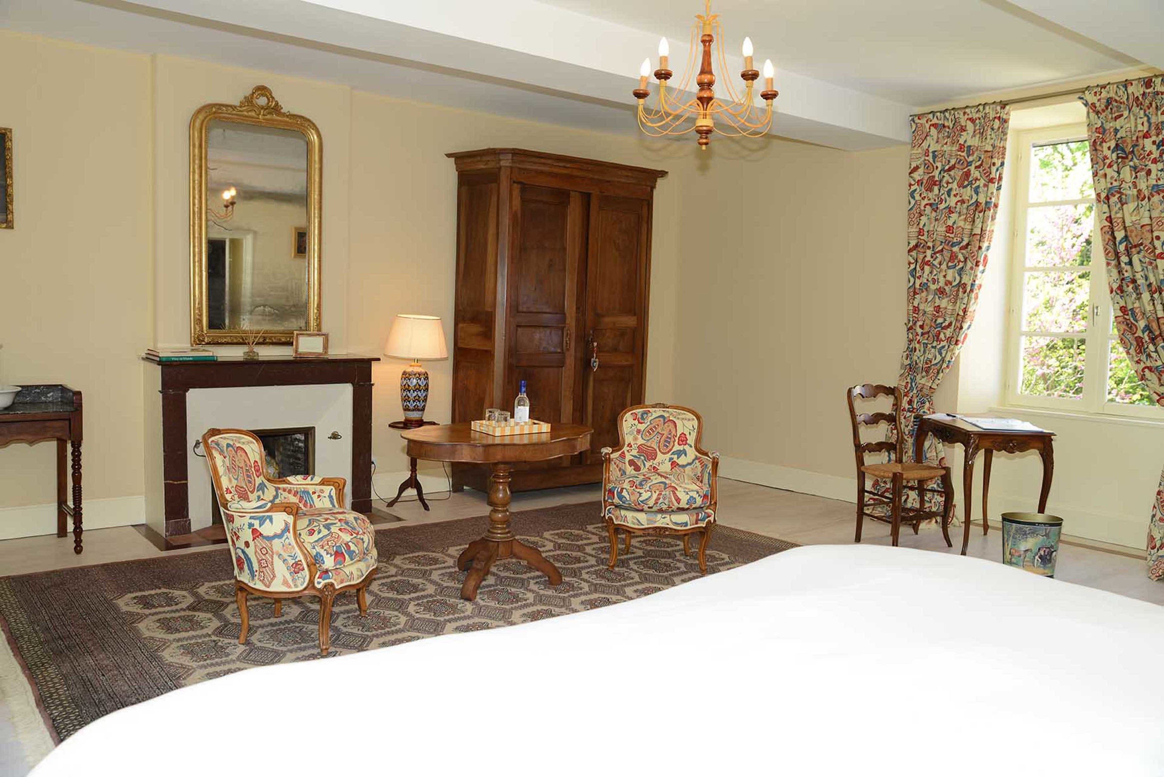 A bedroom with a large bed, two armchairs, a wooden cabinet, a fireplace, and a chandelier.
