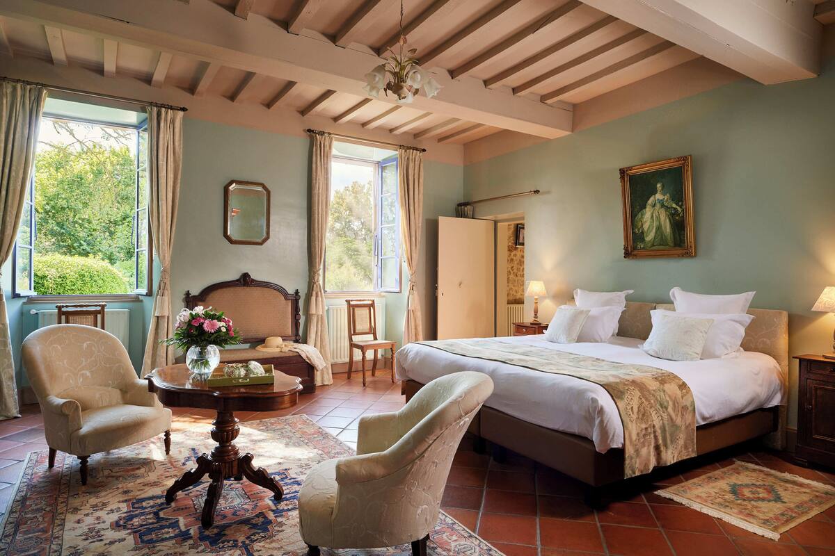 A bedroom with a large bed, a chair, a table with a vase, and a painting on the wall.