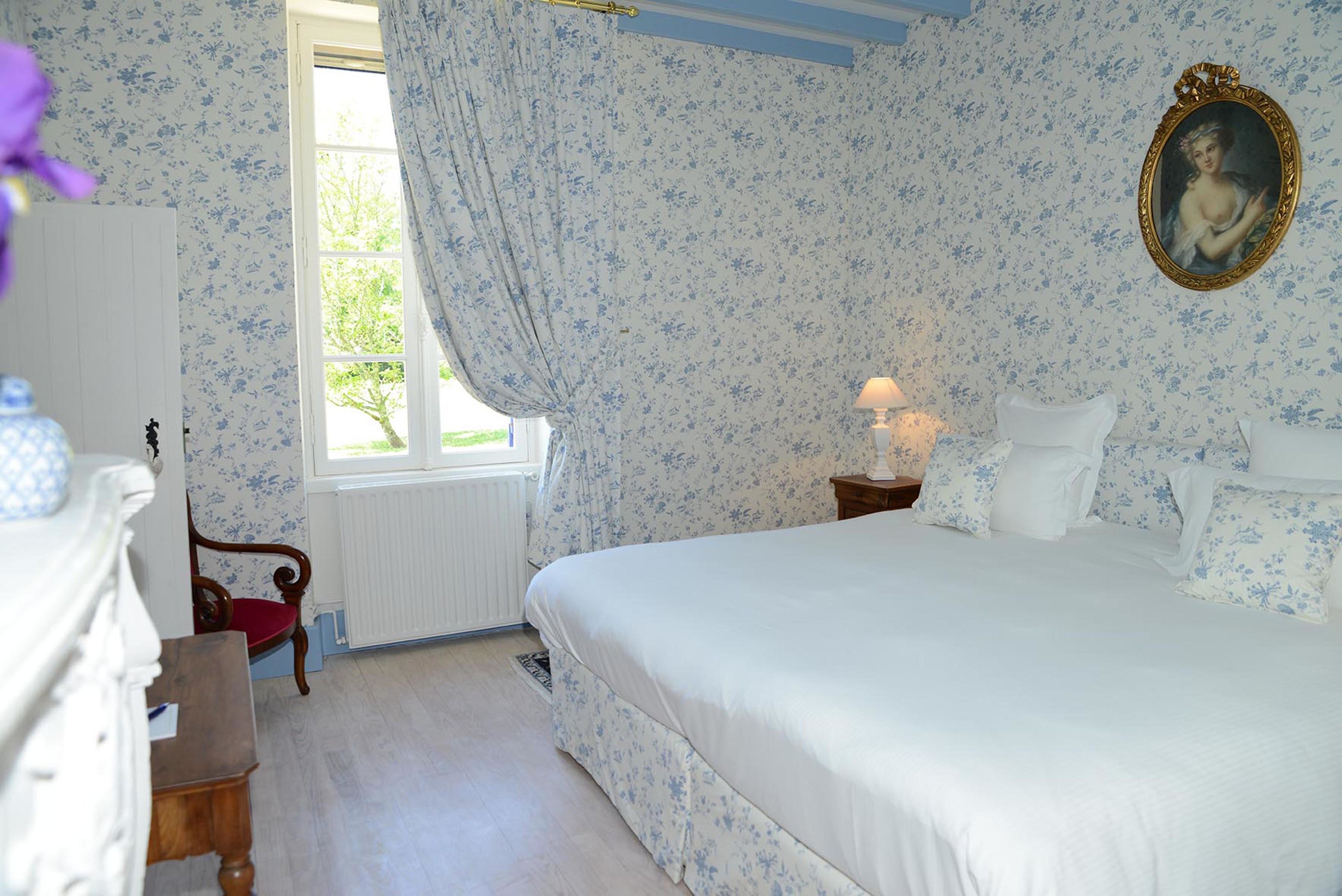 Une chambre à coucher comprenant un lit, une table de chevet, une chaise, une fenêtre avec des rideaux et un mur orné d’un tableau encadré.