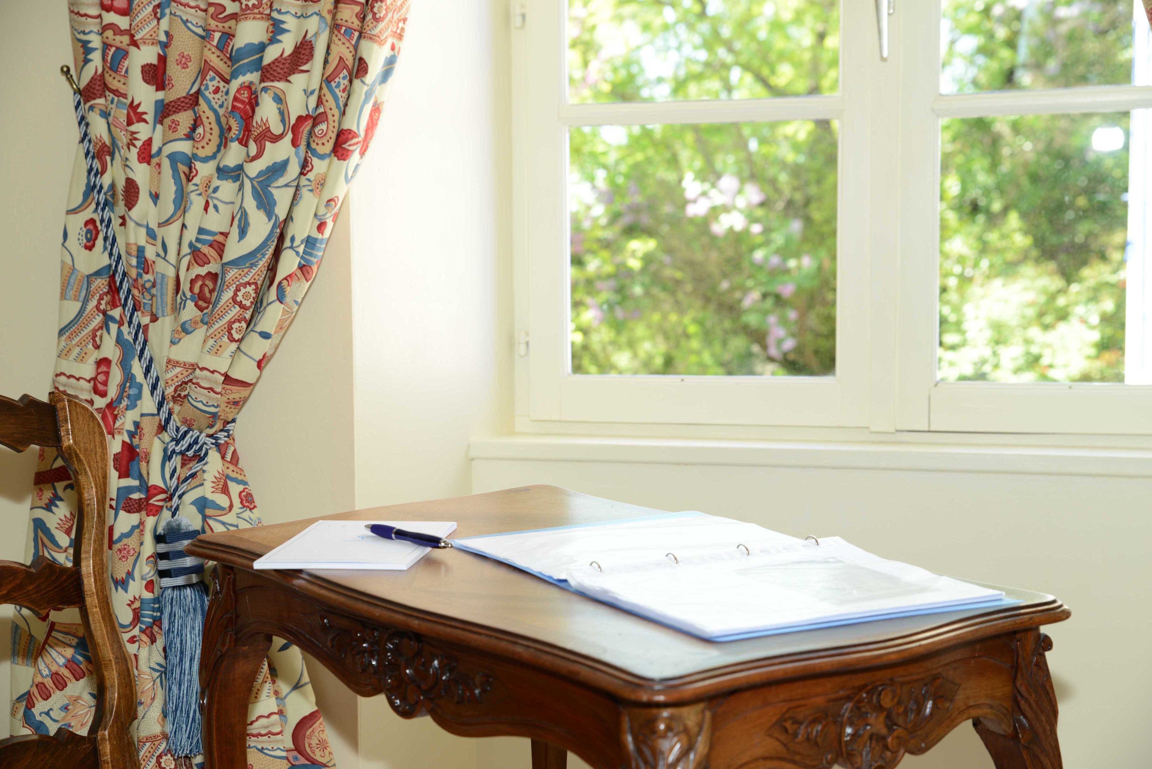 A wooden desk with papers and a pen, a chair, and a window with a view of greenery.