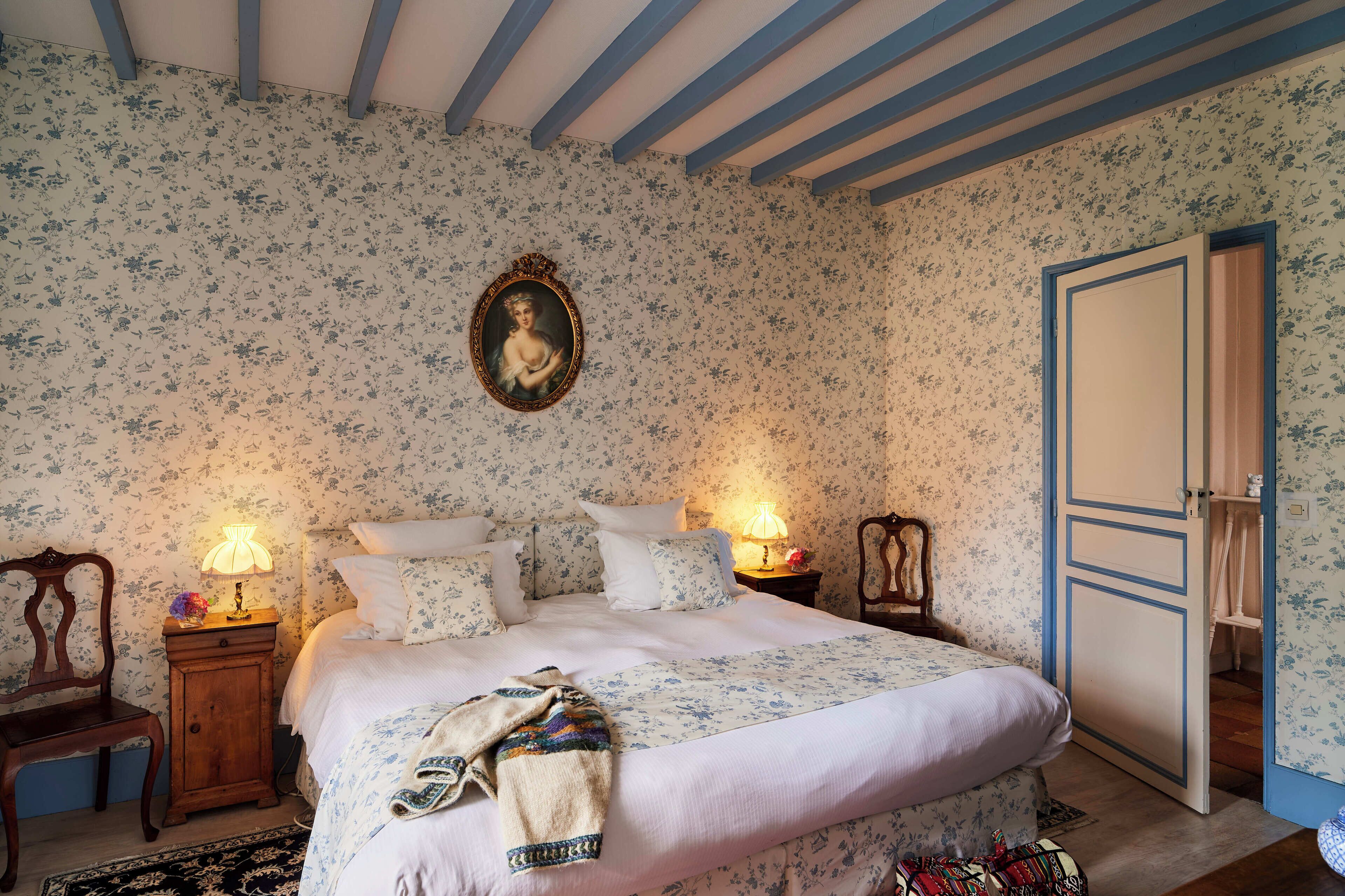 A bedroom with floral wallpaper, a bed with white bedding, two bedside lamps, a wooden nightstand, a chair, and a wardrobe with a blue door.
