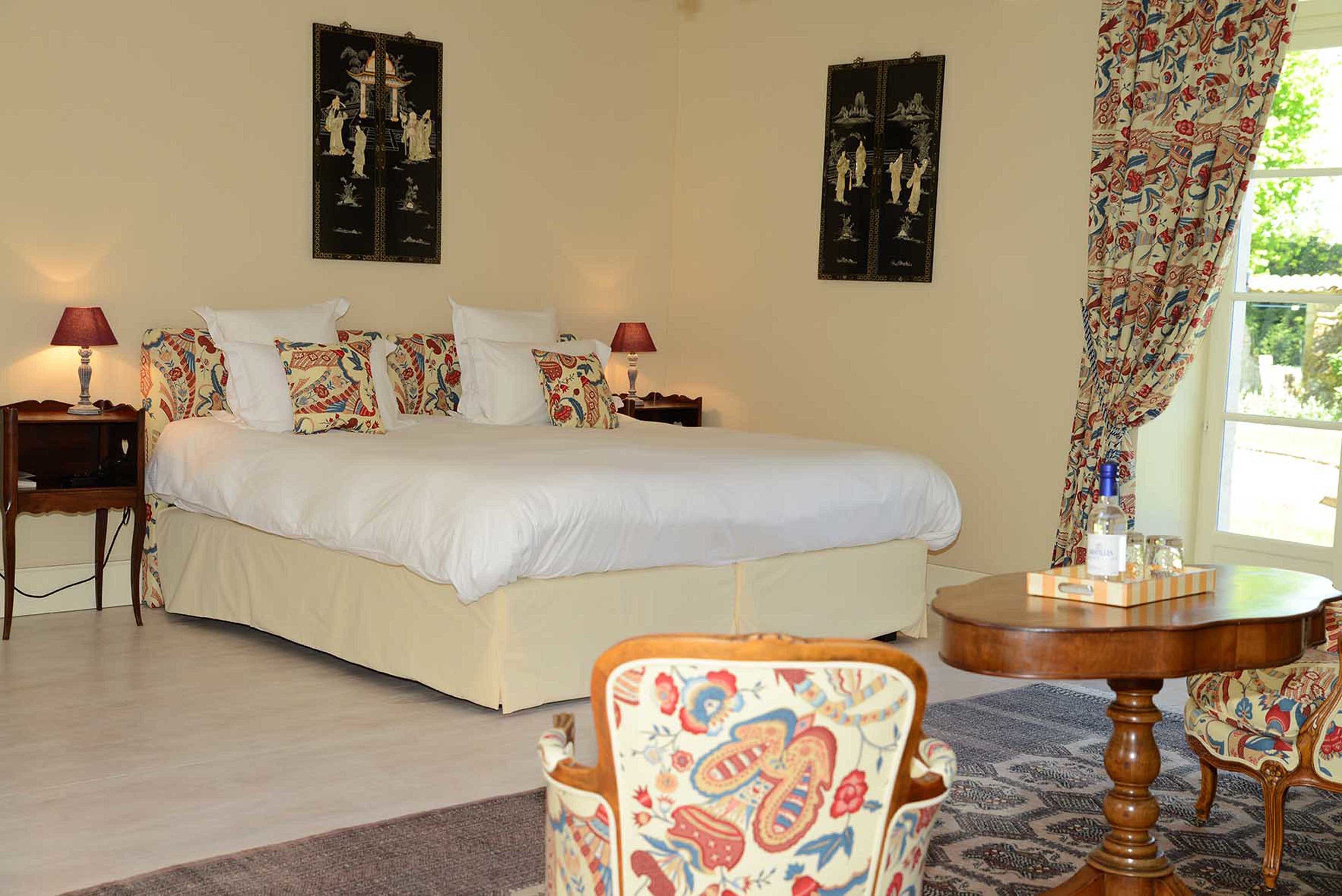 A bedroom with a large bed, two bedside tables with lamps, a round wooden table, and patterned chairs.
