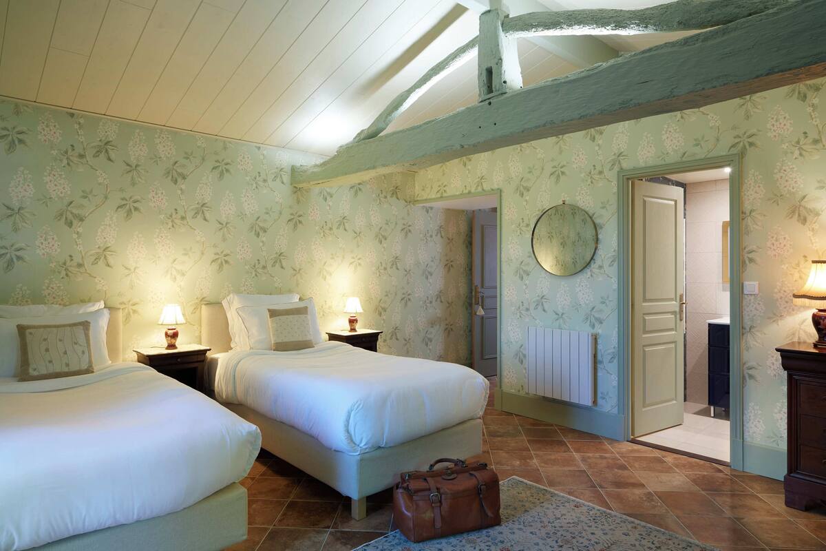 A bedroom with two beds, a wooden ceiling, floral wallpaper, and a door leading to another room.