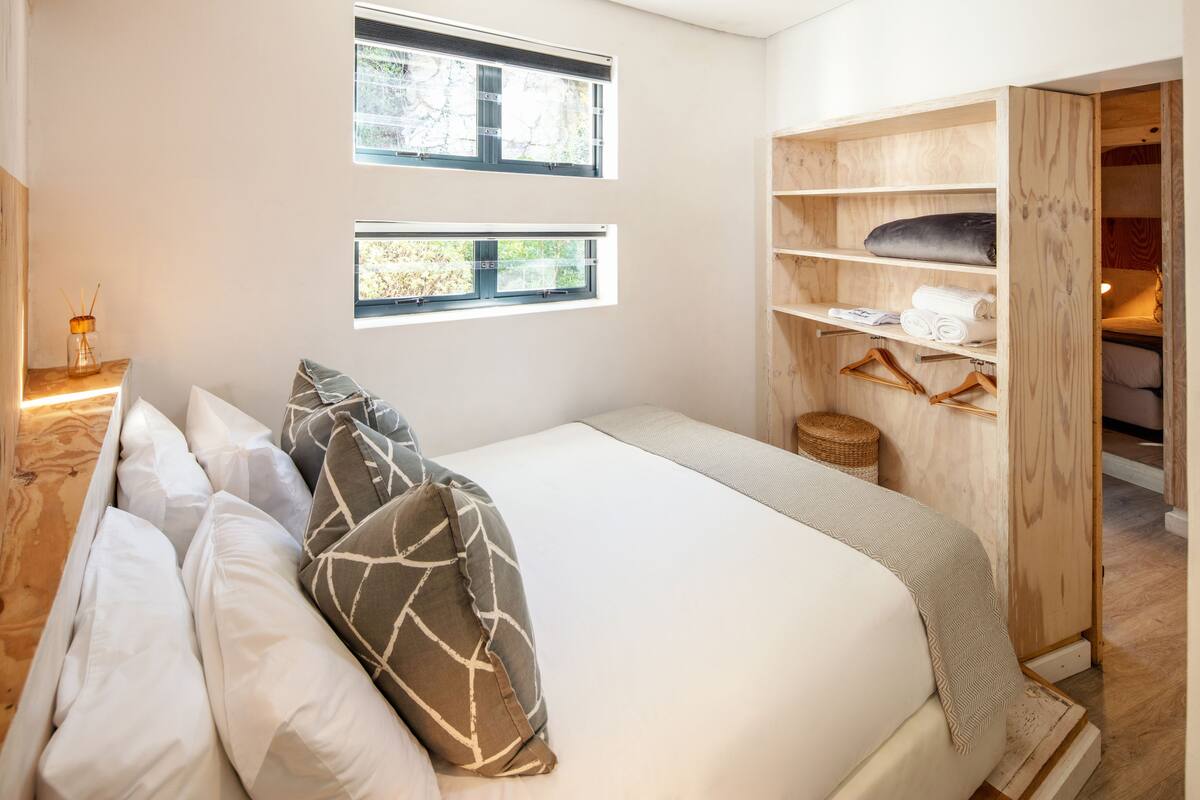 Quarto com cama, duas janelas, guarda-roupa de madeira e uma pequena mesa com abajur.