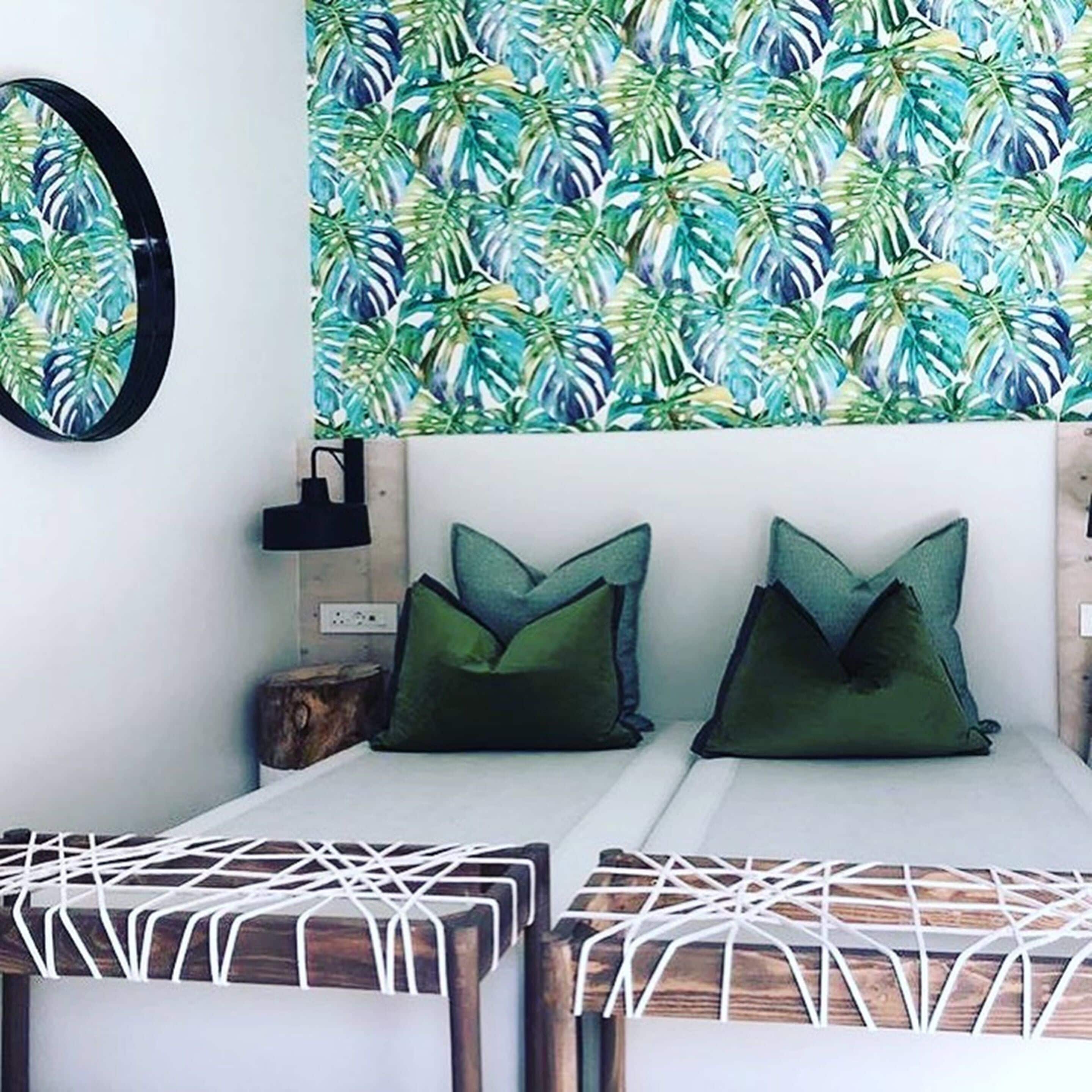 Une chambre à coucher avec un lit, deux oreillers, une table de chevet, une horloge murale et un papier peint orné de motifs de feuilles tropicales.