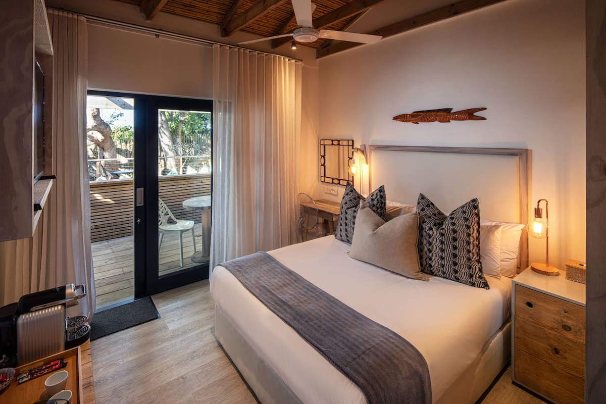 Quarto de hotel moderno com uma cama grande, um ventilador de teto, uma mesinha de cabeceira de madeira e vista para a área externa através de uma porta deslizante.