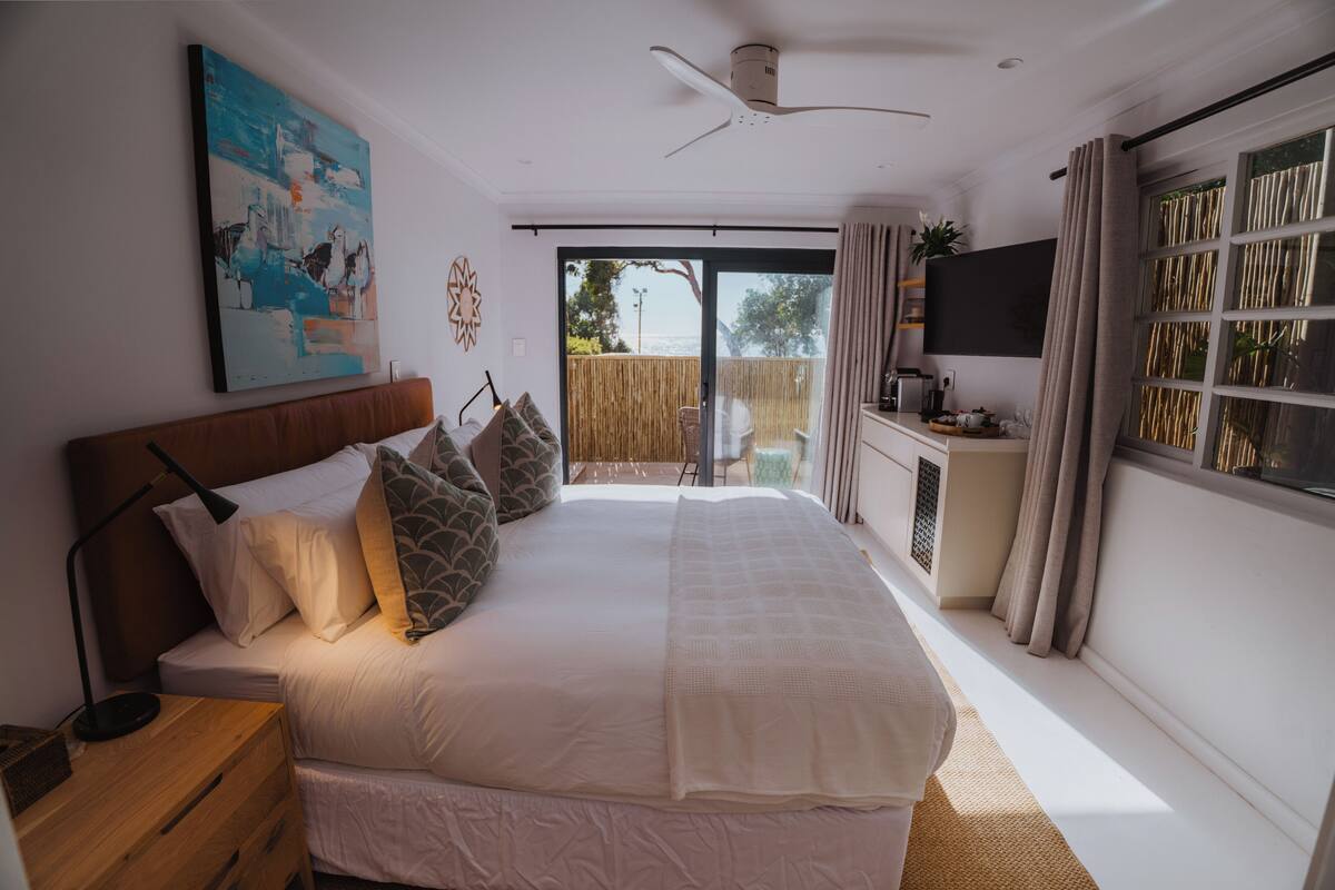 Quarto com uma cama grande, um ventilador de teto, uma TV e uma porta de vidro deslizante para uma varanda.