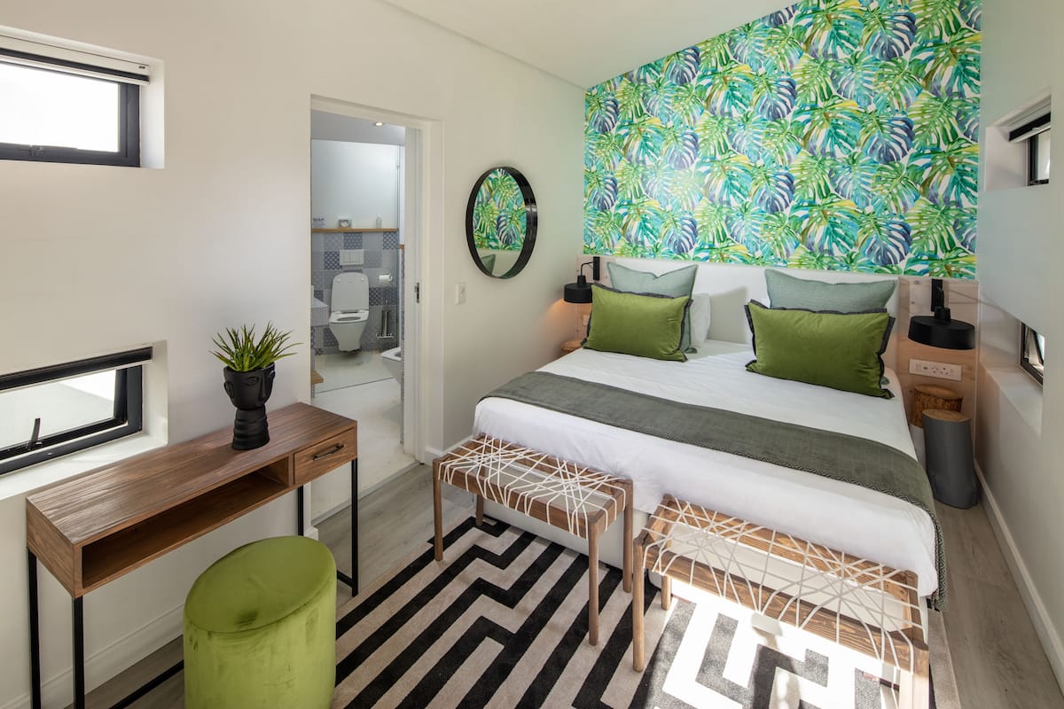 Quarto moderno com cama, mesas de cabeceira, escrivaninha e um banquinho verde.