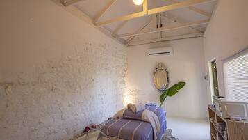 A bedroom with a bed, a wooden ceiling, an air conditioning unit, a mirror, and a window with blinds.