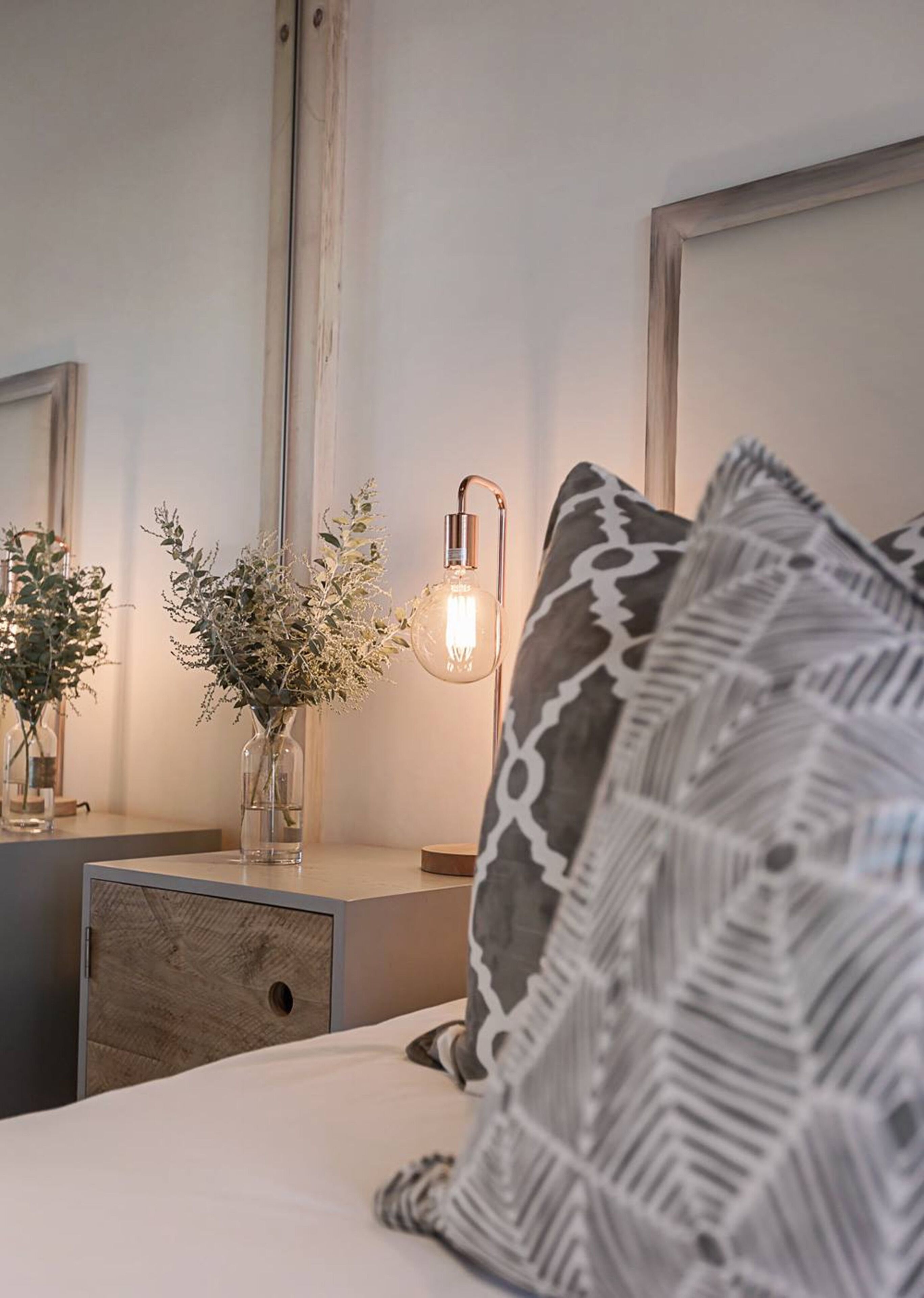 A bedroom with a bed, a bedside table with a vase of dried flowers, a wall-mounted light, and a patterned pillow.
