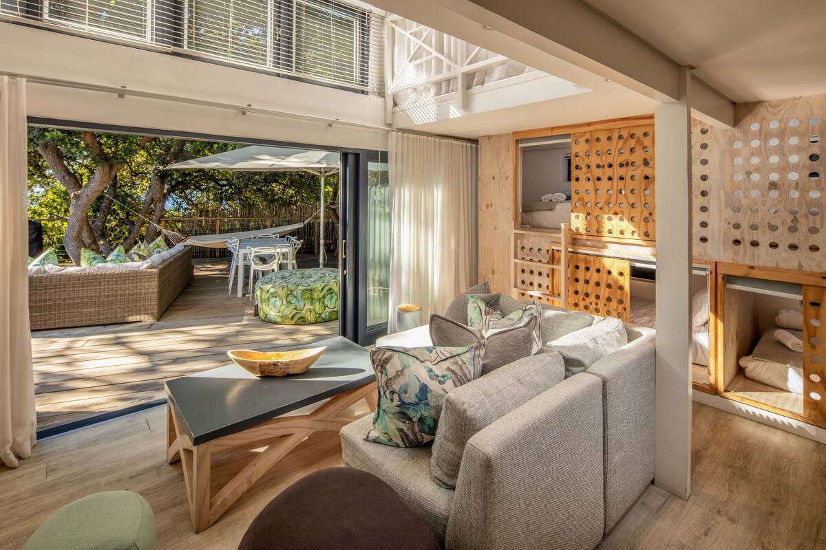 Uma sala de estar moderna com sofá, uma mesa e um armário de madeira. Há uma porta de vidro que dá para uma área externa com uma rede e uma mesa de jantar.