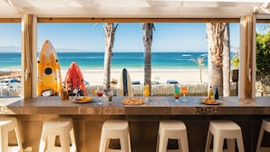A beachside bar with a view of the ocean, featuring kayaks, a surfboard, and a variety of food and drinks on the counter. - The Bungalow  (Plettenberg Bay)